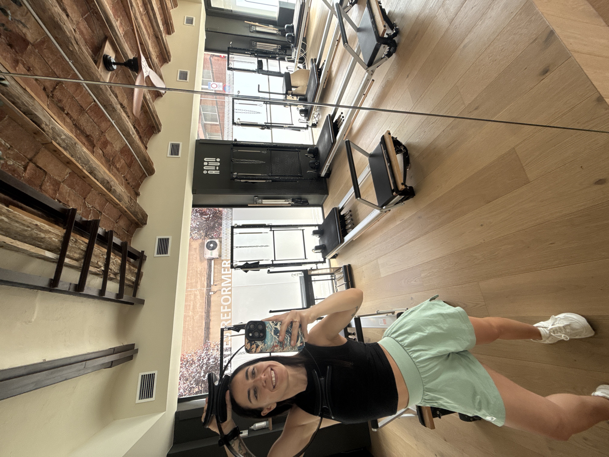 A woman is taking a selfie in a Pilates studio in Barcelona, Spain. She is wearing a black crop top and light green shorts, and holding a Pilates ring above her head. The studio has wooden floors and a high ceiling with exposed beams. There are several Pilates reformers lined up in the room, and a large mirror reflects the scene. The woman is smiling at the camera. The location appears to be a commercial space in an urban setting, given the visible building facades and window reflections.