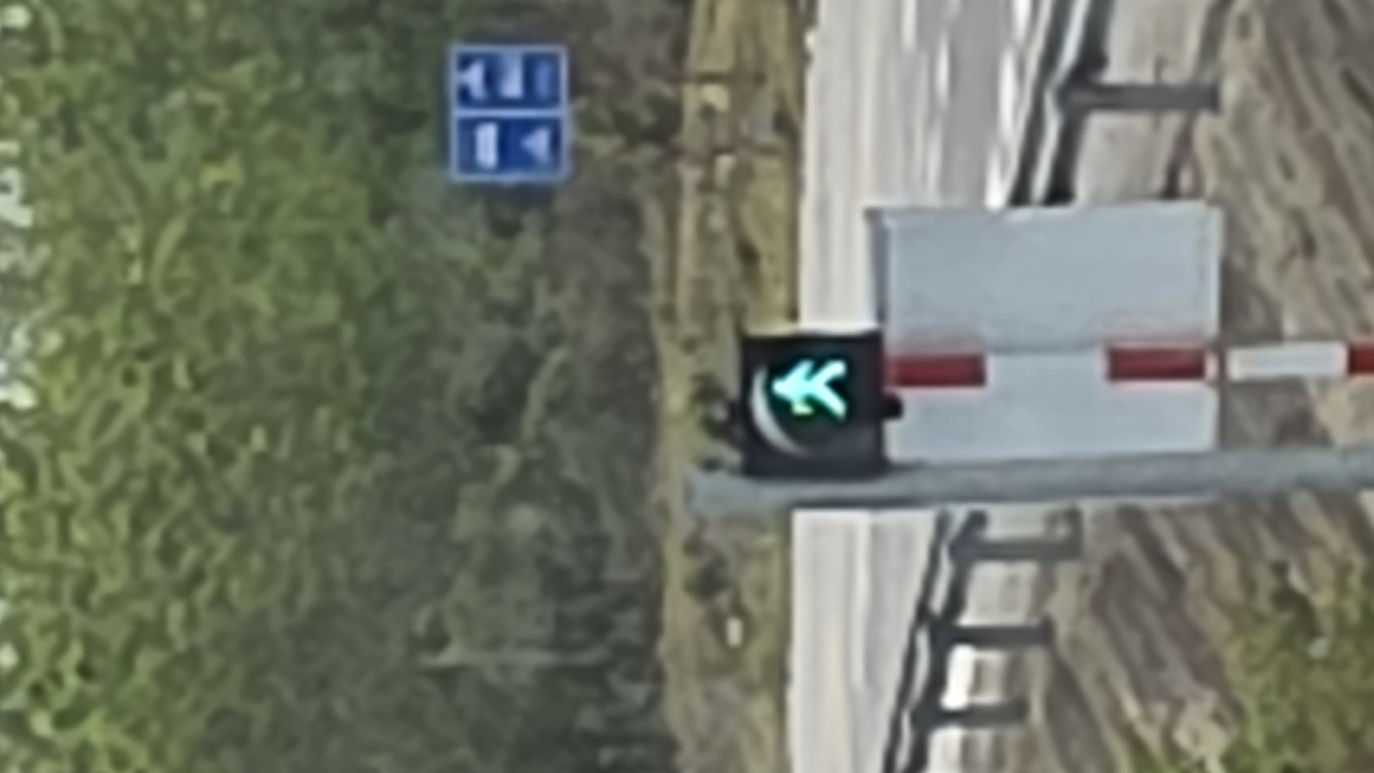 The image shows a railway crossing in Đăk La, Vietnam. A green arrow traffic light is illuminated, indicating permission to proceed. A railway crossing barrier is present, with white and red horizontal stripes. A blue sign with directional arrows is visible in the background. Green foliage is present on the left side of the image. A road surface runs alongside the railway crossing.