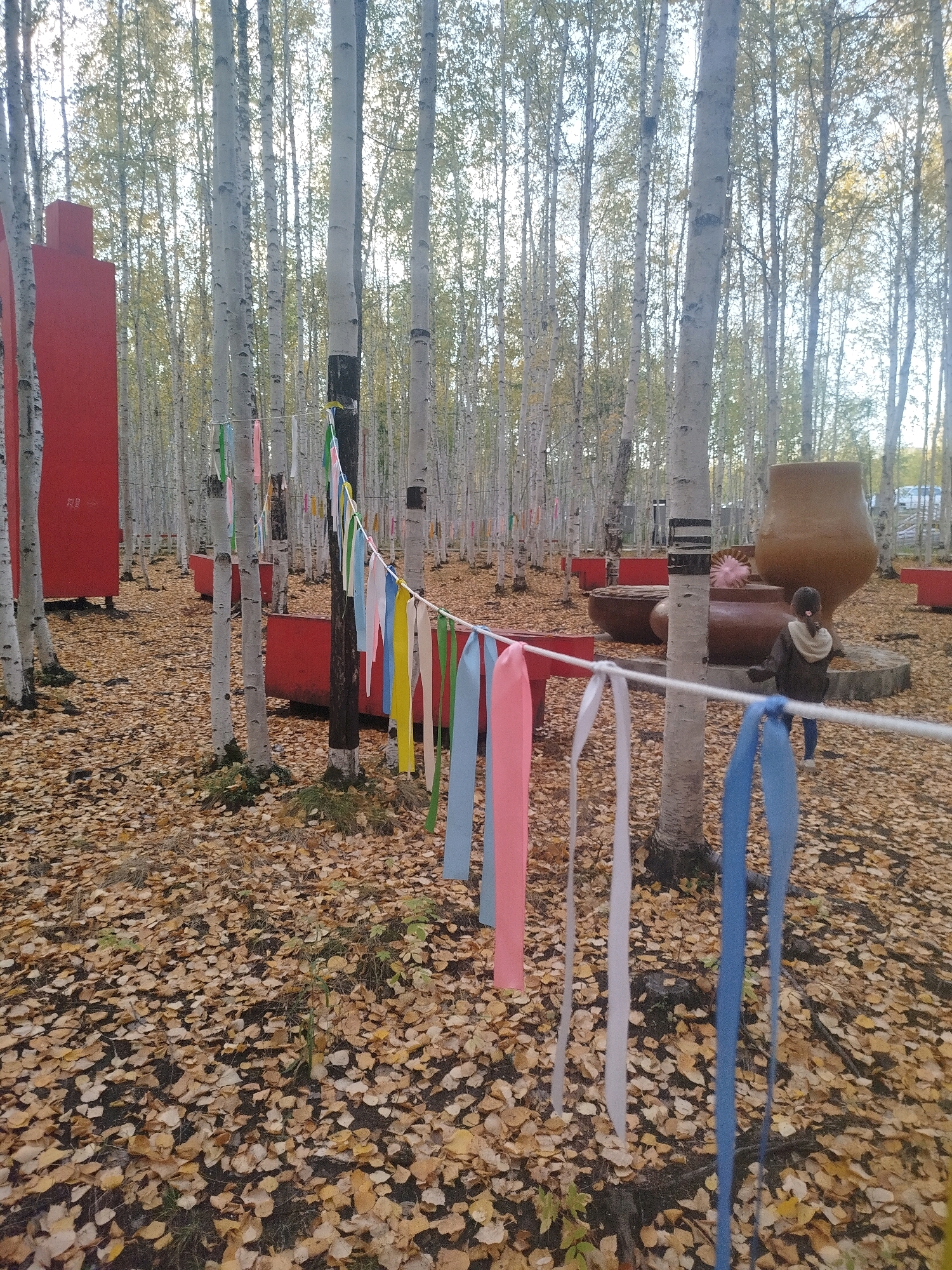 The image depicts an outdoor art installation within a grove of birch trees in the city of Mirny, Russia. The ground is covered in fallen autumn leaves. A string of colorful ribbons, including pink, yellow, white, green, and blue, is stretched horizontally between two trees, with some ribbons dangling down. In the background, to the left, is a tall, rectangular, red structure. To the right, there is a large, tan, ceramic pot, and a young person wearing a dark hooded jacket and jeans is walking away from the camera. Several low, rectangular, red planters are also visible throughout the scene. The overall atmosphere is autumnal and artistic.