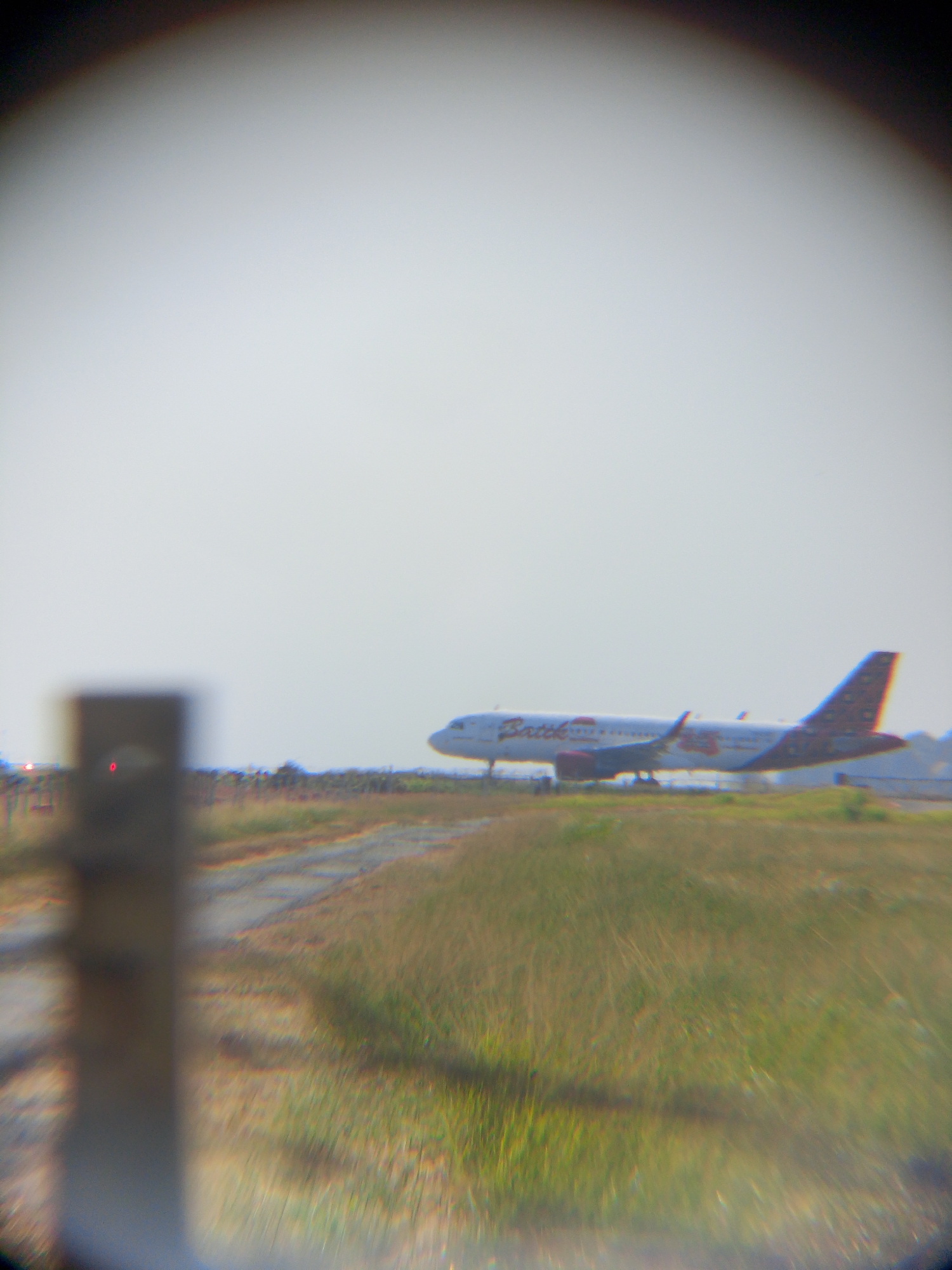 An airplane with "Batik" written on its side is taxiing on a runway at Sultan Aji Muhammad Sulaiman Sepinggan International Airport in Balikpapan, Indonesia. The airplane is white with red and blue markings on its tail. The sky is overcast and the ground is covered with dry grass. A blurred dark object is in the foreground on the left side of the image, possibly a fence post or a piece of equipment. There are no people visible in the image. The overall impression is of a quiet, perhaps early morning, scene at an airport.