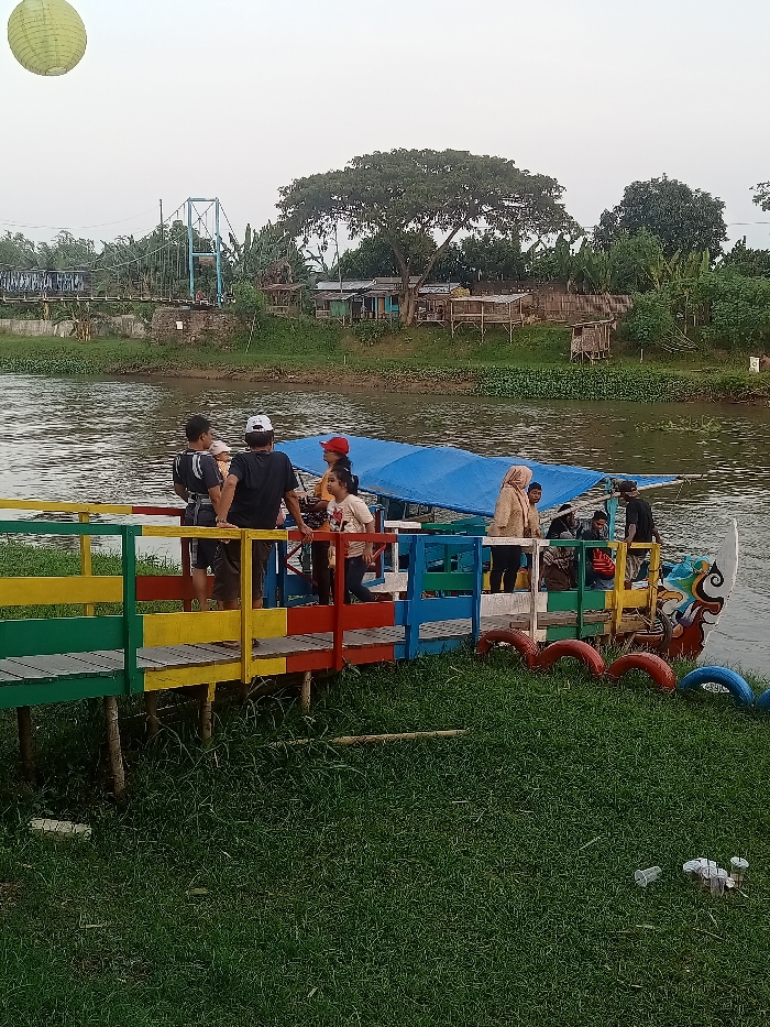 The scene is a riverbank in Jakenan, Indonesia, with a boat docked on a brightly colored, wooden dock. The boat has a blue tarp covering its seats. A group of people, including a woman wearing a hijab, are standing on the dock, interacting with others on the boat. There is a large tree in the background and a footbridge in the distance. The riverbank is covered in green grass and vegetation. The sky is clear. A yellow paper lantern hangs suspended in the air above the scene.