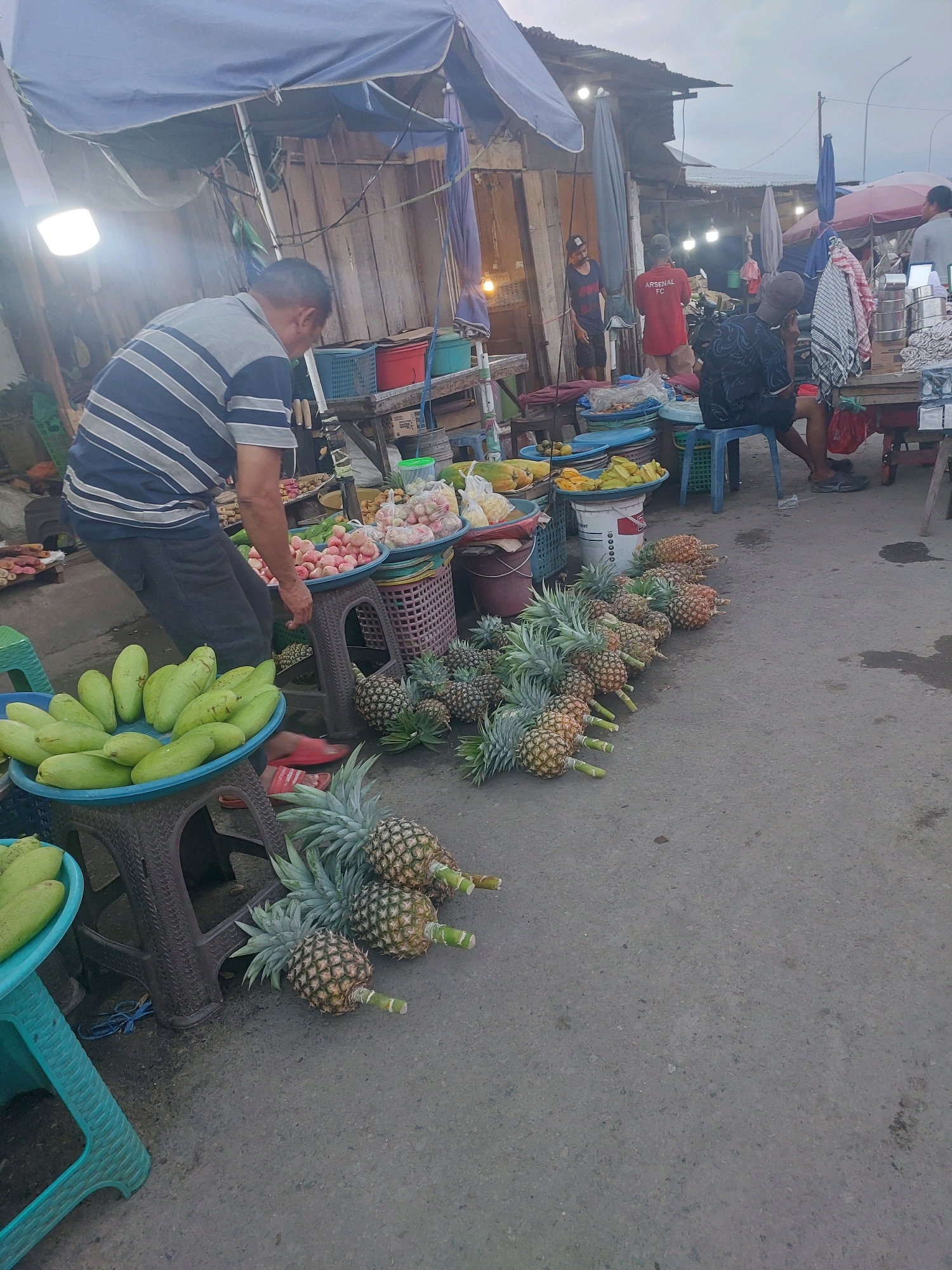 A street market scene unfolds in Ambon, Indonesia, under a pale sky. A man in a striped shirt bends over a display of green fruits, possibly cucumbers or gourds, arranged in a blue plastic basin. Behind him, other market stalls are visible, with awnings providing shade. Several individuals are seen in the background, including one wearing a red shirt with "ARSENAL FC" printed on it, and another seated on a blue stool. Pineapples are prominently displayed, laid out in rows on the asphalt ground. The market appears to be in an outdoor setting, with various fruits and vegetables available for purchase.