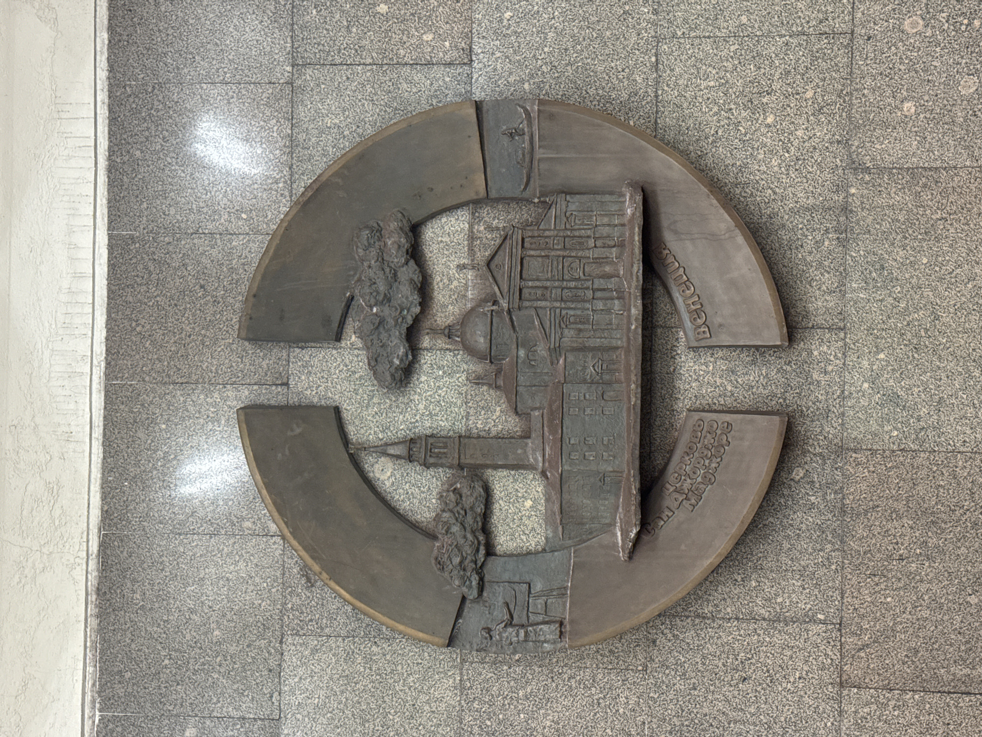 A bronze circular plaque is mounted on a gray tiled wall in Moscow, Russia. The plaque depicts a cityscape with a building featuring a dome and a tower, flanked by trees. Text in Russian is inscribed on the plaque. The wall on which the plaque is mounted features small square tiles. To the left of the wall, there is a white wall.