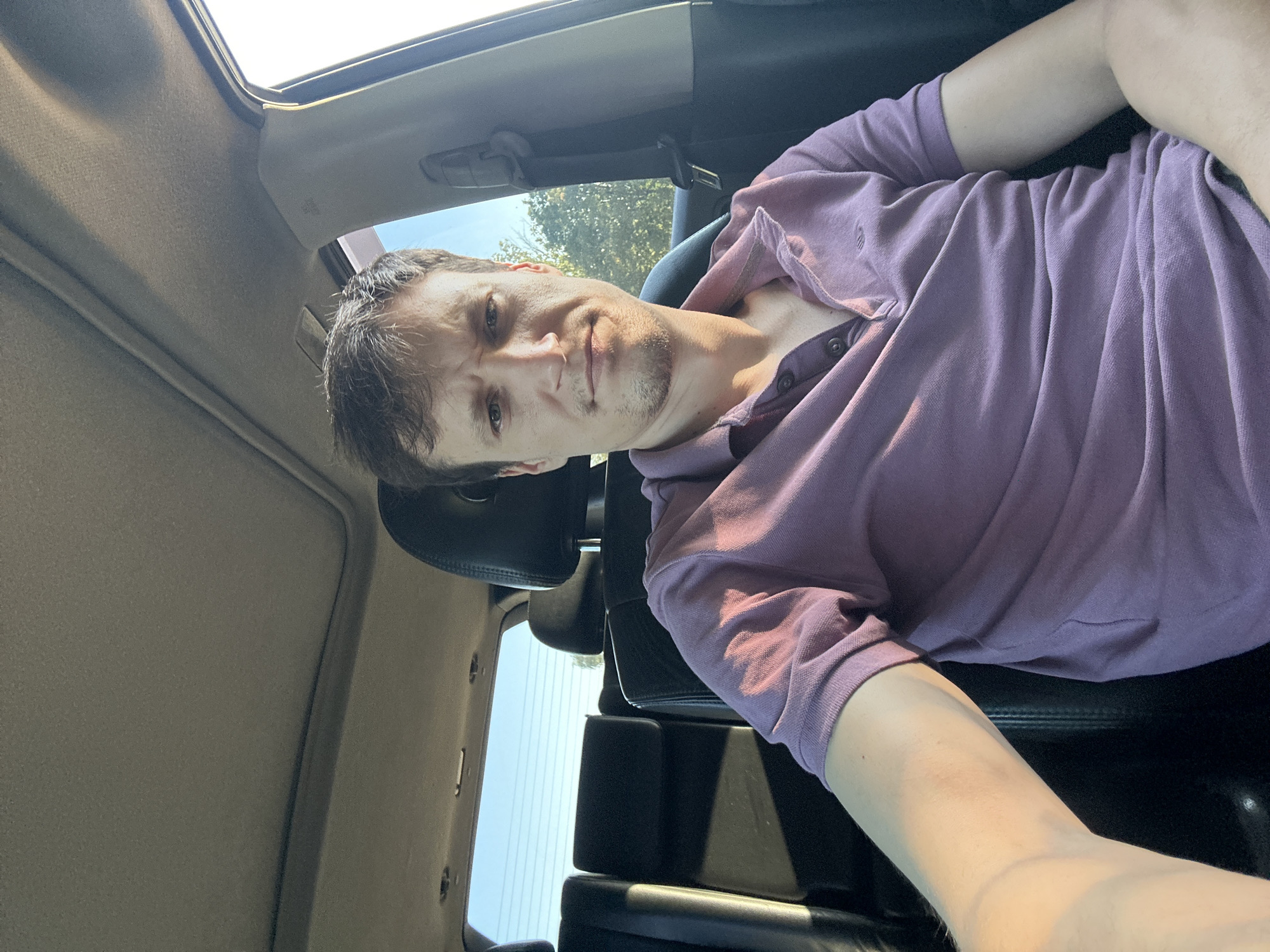 A young Caucasian man with light brown hair and a short beard is sitting in the driver's seat of a car, facing the camera. He is wearing a purple polo shirt. The car interior is visible, including the dashboard, car seats, and a sunroof. The windows are open, revealing a sunny outdoor environment with trees and a clear blue sky. The man appears to be looking directly at the camera with a neutral expression. The image was taken in Makiv, Ukraine.