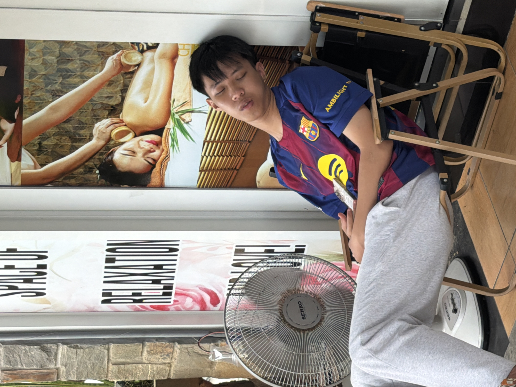 A young Asian man is captured resting or sleeping indoors, leaning against a wall. He is wearing a blue and red FC Barcelona jersey with "AMBILIGHT TV" text on the sleeve, and light grey sweatpants. His eyes are closed, and his mouth is slightly open, indicating a state of deep relaxation or sleep.
Behind the man, a large promotional poster depicts a serene woman enjoying a spa treatment, with her eyes closed and a gentle smile, as hands apply cupping therapy to her upper body. The poster's background features a textured, warm-toned wall. Below this spa image, another poster is partially visible, displaying the vertically stretched words "SPACE OF" and "RELAXATION," confirming the setting's theme.
To the man's right, several wooden chairs with dark fabric seats are stacked. On the floor nearby, a metal-bladed electric fan and a white bathroom scale are visible, both bearing the brand name "SENKO."
The image conveys a tranquil atmosphere, likely within a spa, massage parlor, or a similar wellness establishment in Đà Nẵng, Vietnam. The time of day is not apparent, but the indoor setting suggests a moment of repose.