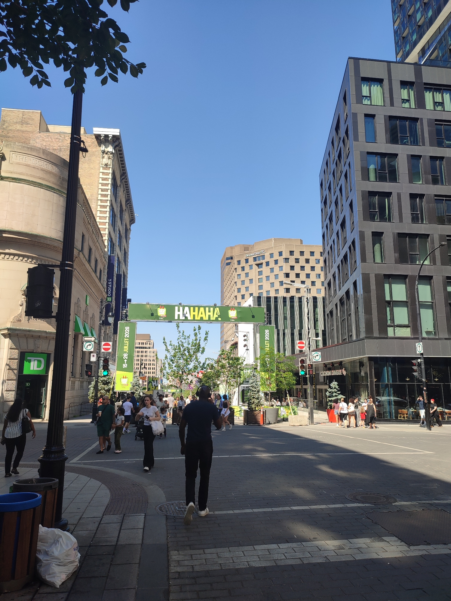 The image captures a sunny day on a city street in Montreal, Canada, likely Sainte-Catherine Street, given the prominent "HAHAHA" banner, a common fixture during Montreal's Just for Laughs festival. A crowd of people are walking and interacting along a pedestrianized area with shops and businesses lining the street. Modern architecture, including tall, glass-fronted buildings, stands alongside older, more ornate structures. Traffic lights and street signs indicate ongoing urban activity. The overall atmosphere suggests a vibrant, bustling urban environment during a public event or busy shopping day.