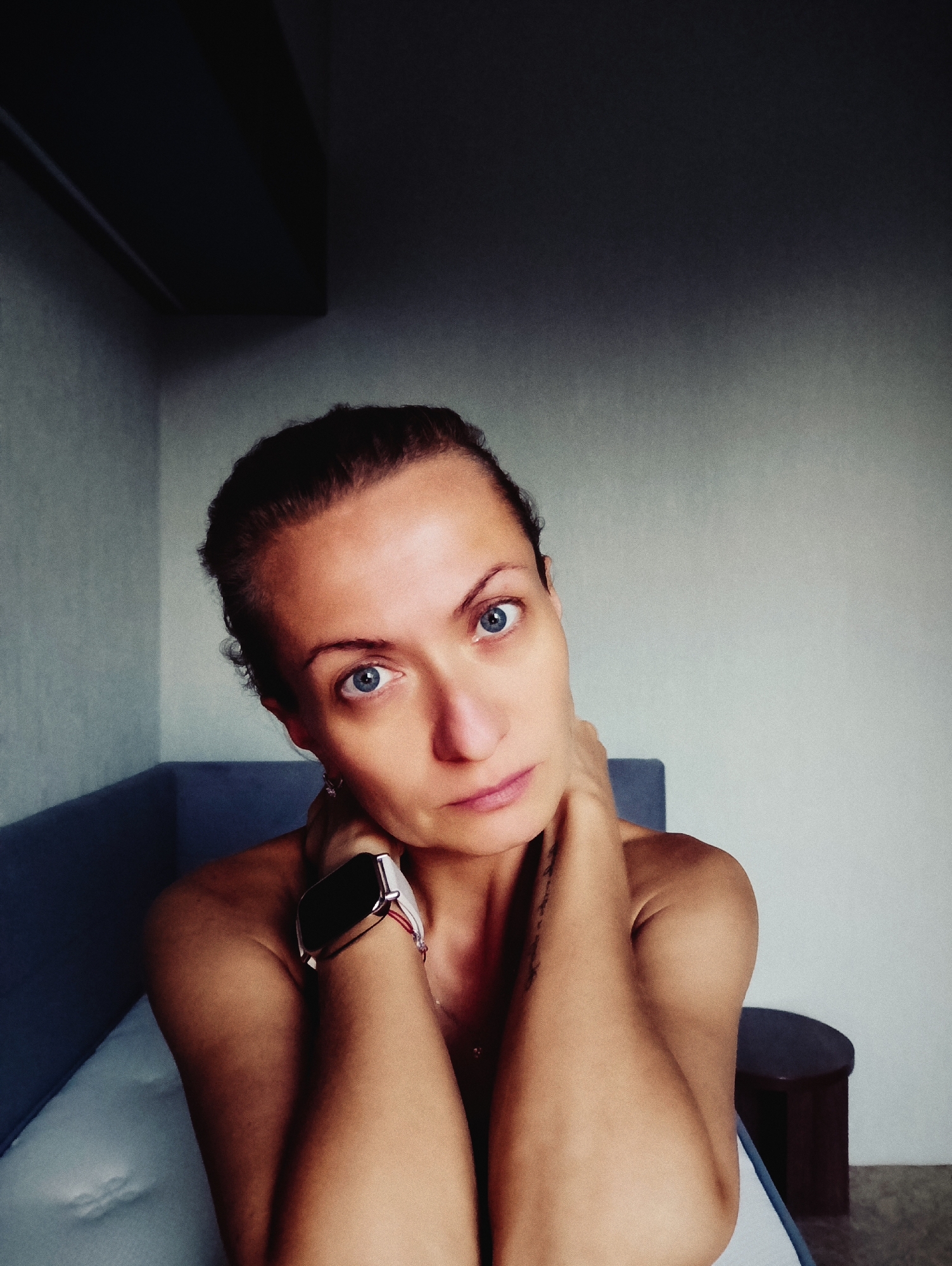 A medium-shot, portrait of a woman with dark hair pulled back, and piercing blue eyes, is shown from the chest up. She is looking directly at the camera with a serious expression. Her hands are clasped behind her neck, and she is wearing a smartwatch on her left wrist. The woman is seated on a light-colored cushion, with a dark blue couch behind her. The background is a wall that transitions from a dark gray in the upper left corner to a lighter gray on the right. There is a dark cylindrical object to the right of the woman, possibly a stool or table. The overall lighting is dim, with shadows casting from the left. The location is not identifiable as Moscow, Russia.