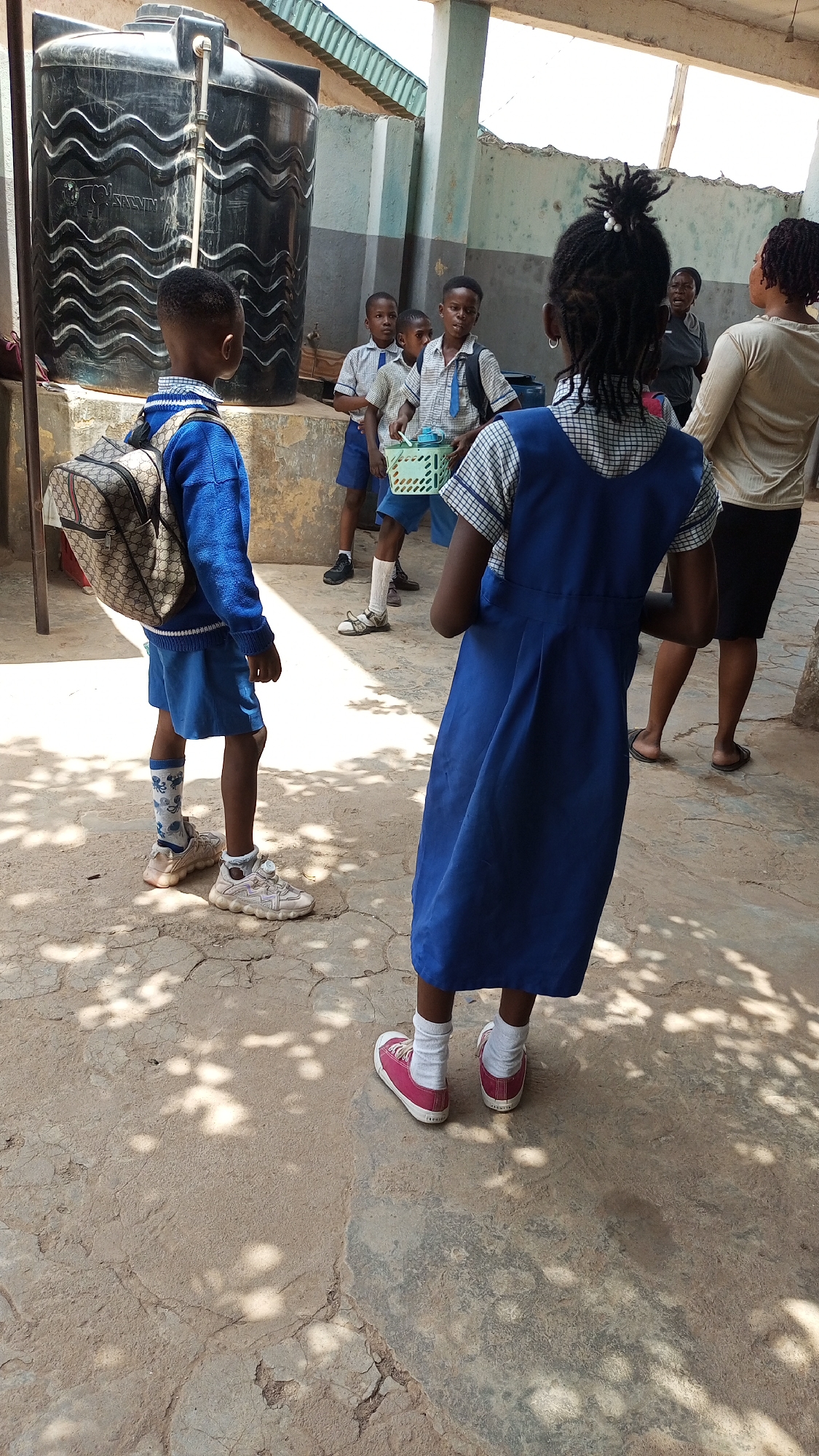 The image depicts a group of people, mostly children in school uniforms, in what appears to be an outdoor courtyard at or near a school in Sabon Tasha, Nigeria. The scene is lit by sunlight casting shadows on the ground.
Several school children are present, dressed in different variations of school uniforms. A young boy wears a blue sweater and skirt with a backpack. Another child wears a blue dress and red shoes. Three boys are visible in short-sleeved, collared shirts and shorts, with one carrying a green plastic basket.
Two adults stand near the children. The taller of the adults, presumably teachers, wears a light-colored blouse and dark skirt.
A large, black water tank is visible in the background. The walls of the courtyard are painted two tones, gray and white. The ground consists of cracked concrete.