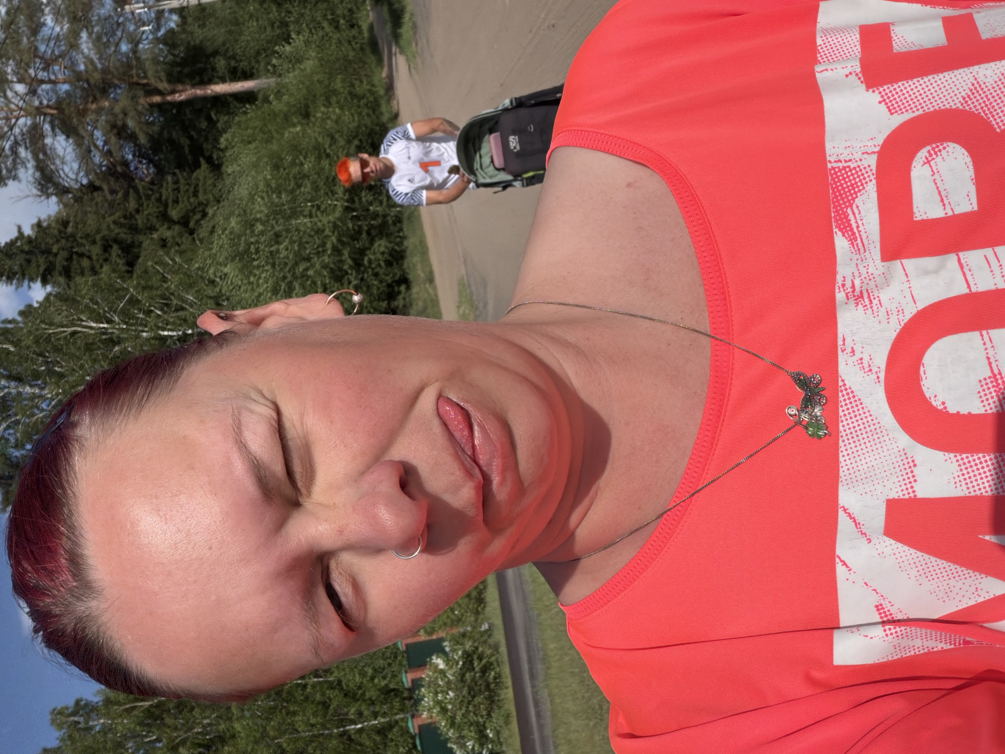 A selfie shows a woman with closed eyes, sticking out her tongue. She has short, reddish hair and is wearing a coral-colored tank top with large white lettering, along with a necklace. In the background, a man with reddish-orange hair is pushing a stroller on a path lined with trees and grass. The image appears to have been taken in a public park or green space. The location is Chaikovsky, Russia.