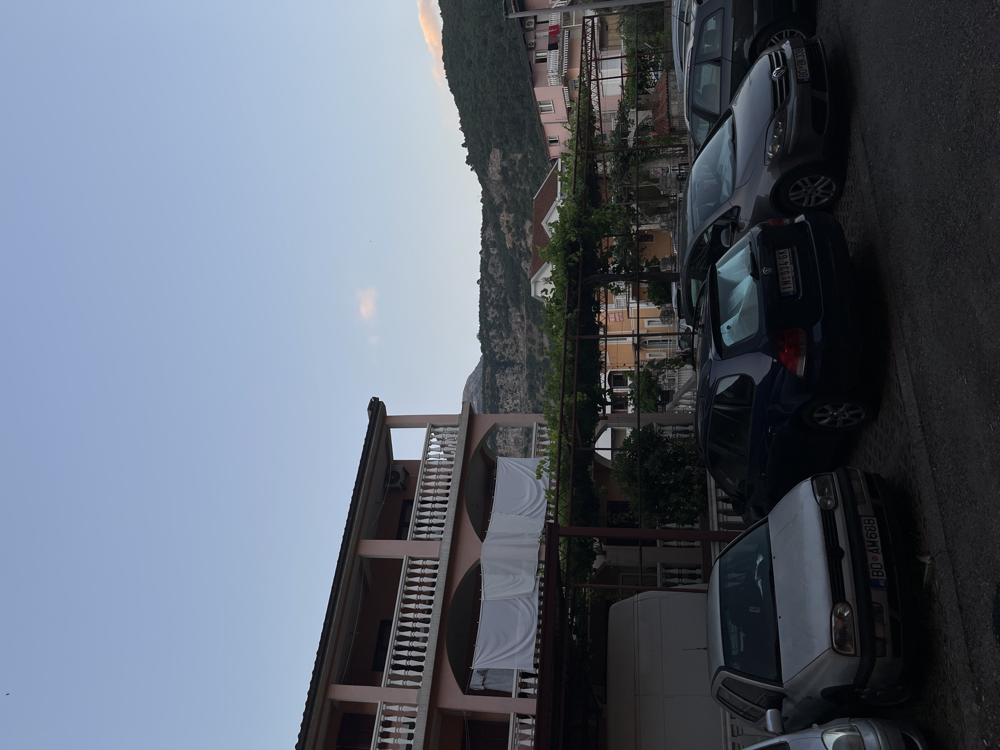 The image depicts a street scene in Budva, Montenegro, with several parked cars in the foreground. A light pink building with multiple balconies and white railings dominates the middle ground. White sheets are hung on a line between columns on the lower level of the building. Beyond the building, a forested mountain slope rises under a pale blue sky with a hint of pink cloud. The parked vehicles include a silver Volkswagen Golf, a dark blue hatchback, and a grey compact car. The ground is paved.