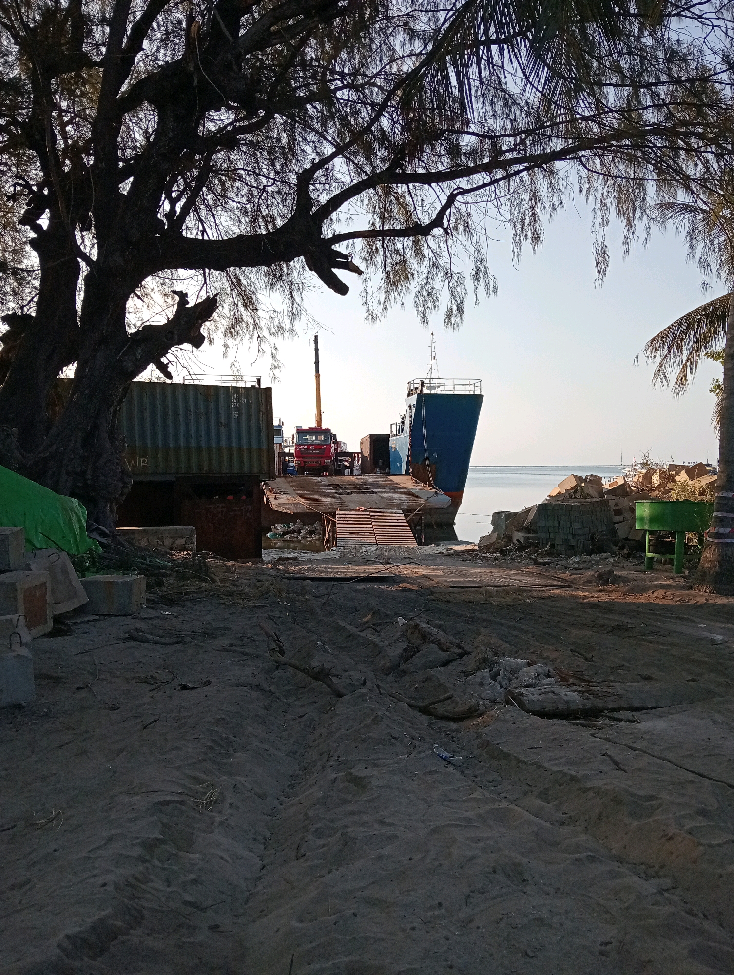 A cargo ship is docked at a pier in Pagerungan Kecil, Indonesia. A red crane truck is positioned on the pier, with its boom extended over a large shipping container. Debris and construction materials are scattered around the pier. The scene is set against a backdrop of the ocean and a clear sky. The time of day appears to be late afternoon, with the sun casting long shadows. The area seems to be undergoing some form of construction or renovation.