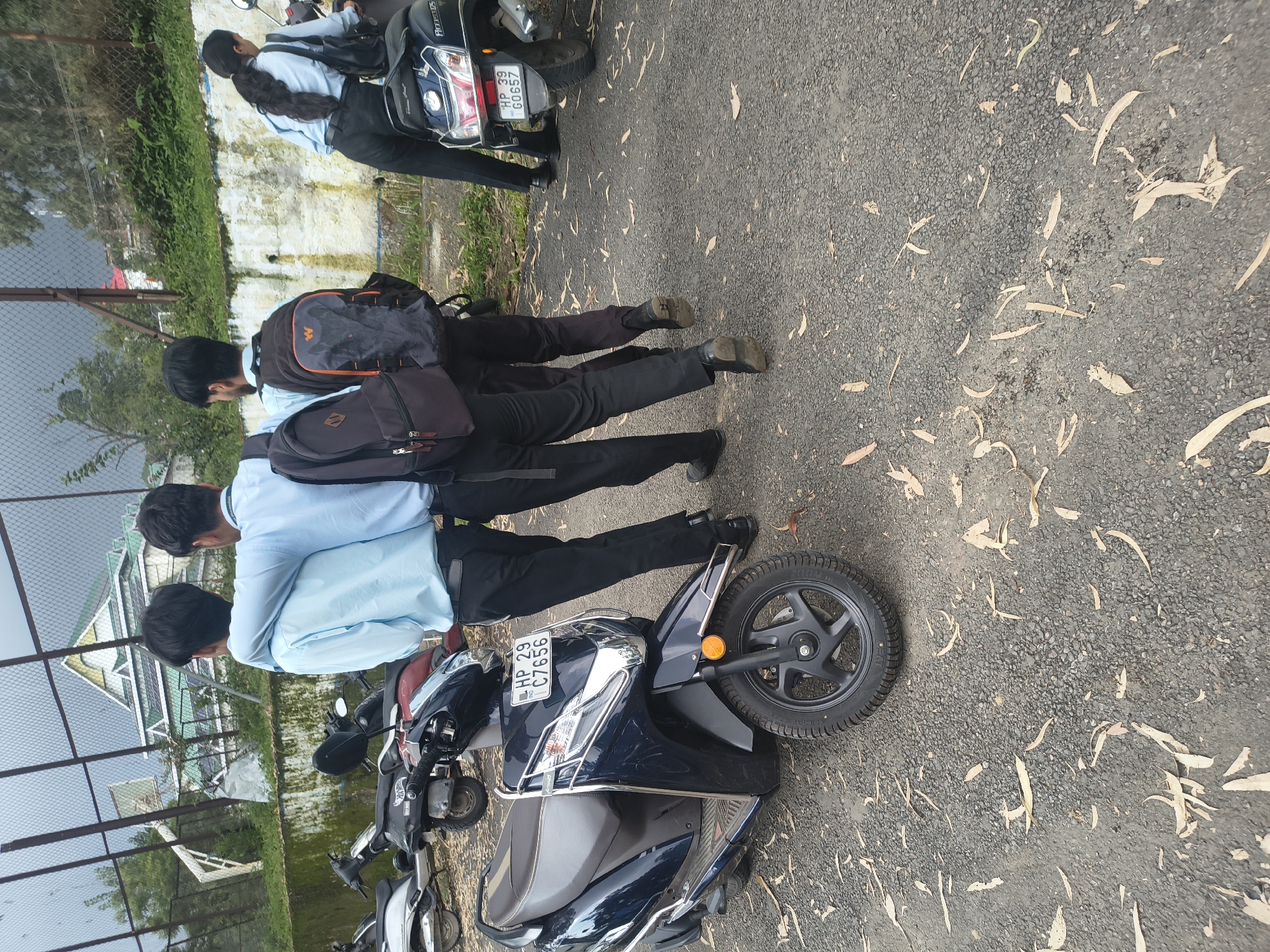 Three young men, two with their arms around each other, walk away from the camera in what appears to be a university campus in Dharamshala, India. They are dressed in light blue shirts and dark pants, and two of them are carrying backpacks. The ground is paved with asphalt and covered in fallen leaves. In the background, a metal fence encloses a basketball court, and beyond that, a modern building with a green roof is visible. Several parked scooters are also present in the scene, one of which has a license plate that reads "HP 29 C7656". A fourth individual, a young woman wearing a light blue shirt and dark pants, is seen from behind, mounted on a scooter to the right of the frame. She is also carrying a backpack and has her hair tied in a ponytail. The overall atmosphere is casual and suggests a group of students on a campus.