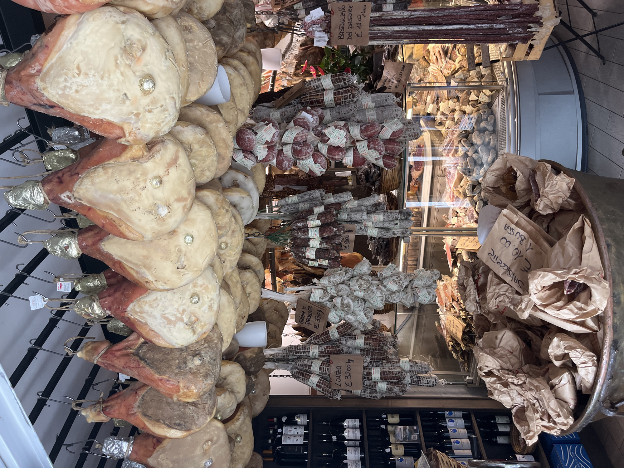 The image depicts a butcher shop in Nemi, Italy, showcasing a wide assortment of cured meats. Numerous whole cured hams, some with gold foil-wrapped tips, hang from the ceiling, obscuring the upper portion of the shop. Below these, strings of various types of sausages and cured salumi are suspended. Some of these sausages are labeled with prices, such as "Lonzo €32.00/kg" and "Salame di Prosciutto €33.00/kg."
In the foreground, a large, rustic copper pot or vat is filled with crumpled brown paper bags. Some of these bags appear to contain pieces of meat. One visible label on a bag reads "SALICCETTE €10.00 BUSA."
To the left of the hanging meats, a tall wooden shelf unit is stocked with several rows of wine bottles.
The background features a refrigerated display case filled with more cured meats, sausages, and what appear to be cuts of pork. The shop has tiled flooring. The overall impression is of a well-stocked, traditional Italian butcher shop specializing in cured meats. There are no people visible in the image.
