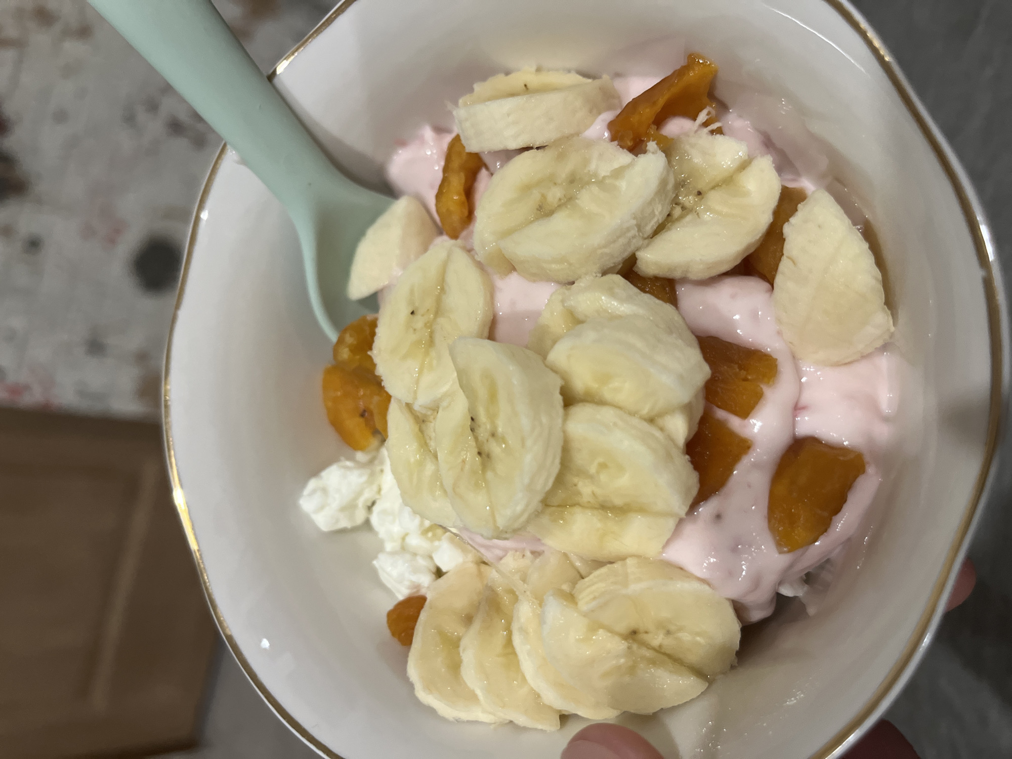 A white bowl with a gold rim is filled with a mix of food items. There are slices of banana, pieces of dried apricot, pink yogurt, and cottage cheese visible. A light green spoon rests on the edge of the bowl. The bowl appears to be held or resting near a light-colored surface. The setting is indistinct, providing no specific evidence of location in Chaikovsky, Russia.