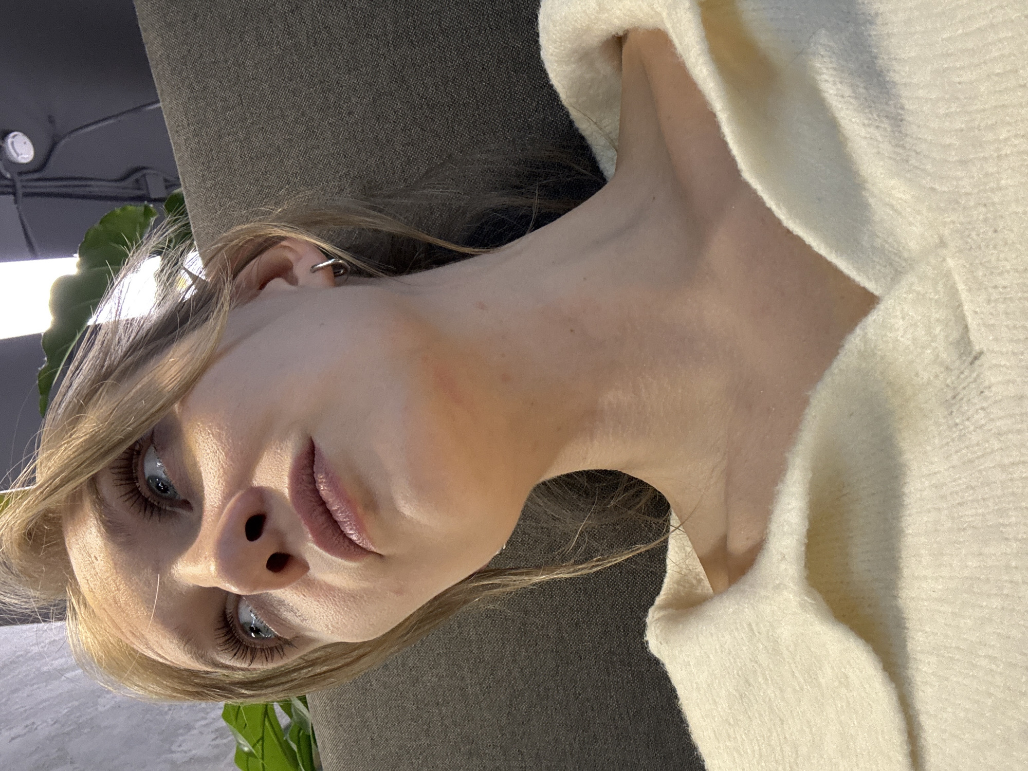 A fair-skinned young woman is lying down, her head tilted toward the viewer. She has blonde hair, blue eyes, a piercing in her ear, and pink lipstick. She is wearing a white sweater. The background is a grey fabric, likely a couch or similar furniture, and a glimpse of a green plant and a ceiling with lighting fixtures is visible in the upper left corner. There is no indication of the location being Mozhvai, Russia in the image.
