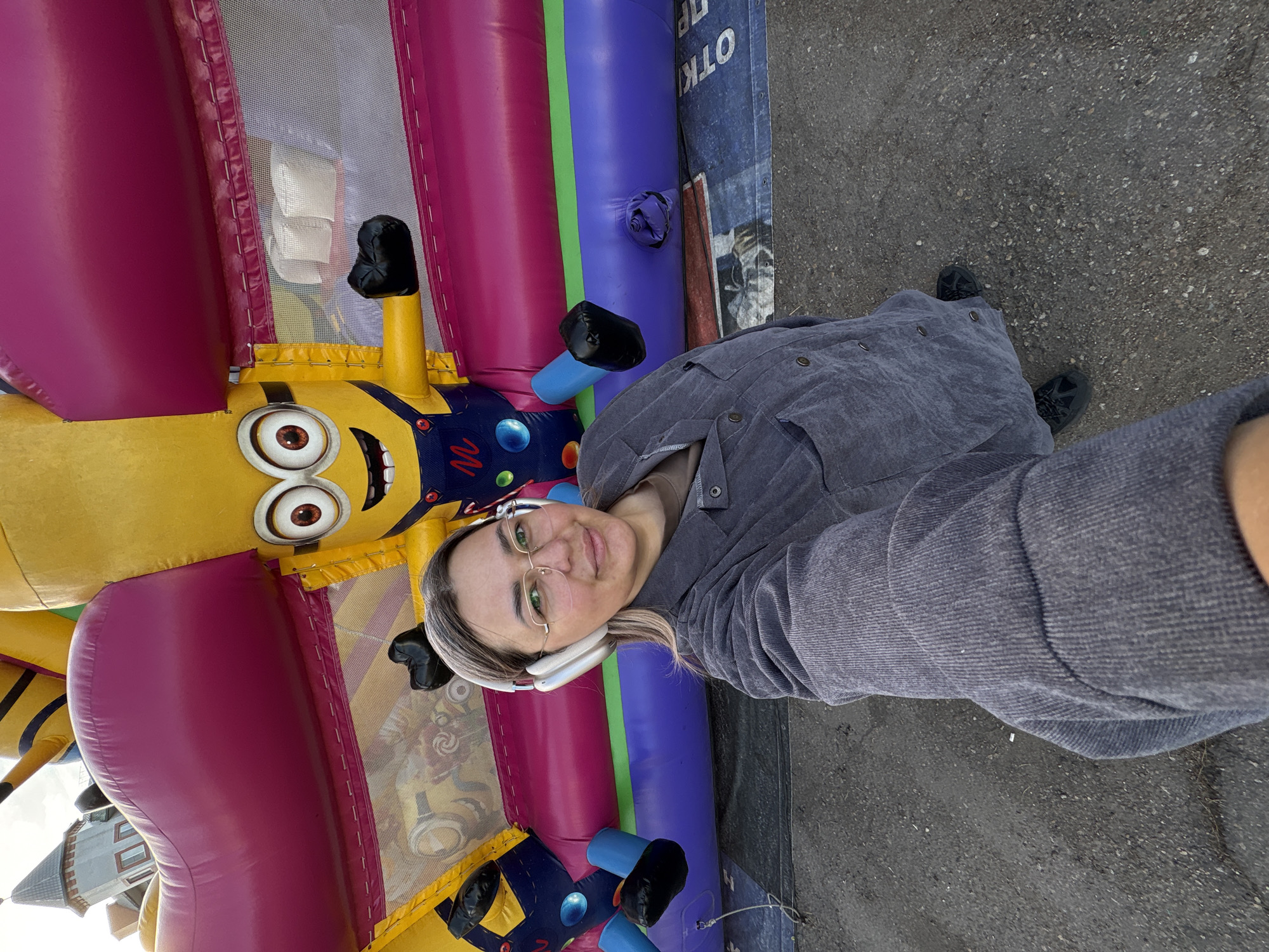 A selfie photograph depicts a woman standing in front of an inflated play structure. The woman is wearing eyeglasses, headphones, and a gray button-down shirt. She has light-colored hair. The play structure is brightly colored with inflatable sections of pink, blue, and yellow and features a Minion character. The ground appears to be asphalt. The location in Birobidzhan, Russia is not discernible from the image.