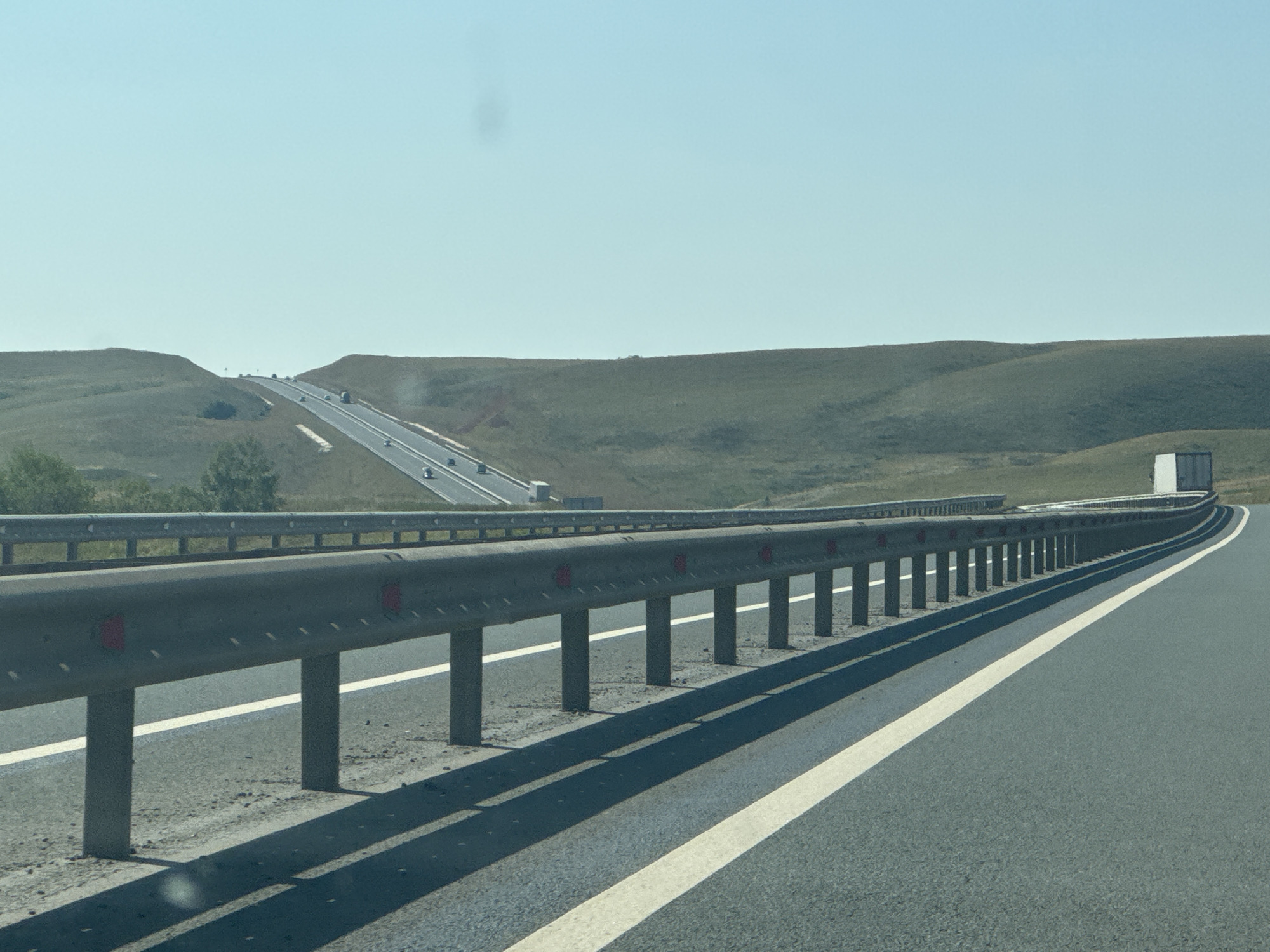 The image depicts a clear sky over a vast, grassy landscape. A multi-lane highway with white lane markings cuts through the scene. On the right side of the highway, a metal guardrail runs parallel to the road. The road curves to the right in the distance, where a white semi-trailer truck is visible. On the left side of the highway, the terrain slopes upwards, covered in dry grass and some scattered bushes. Further in the distance, the highway appears to ascend a hill, with several vehicles visible on it. The overall impression is of a sparsely populated, rural area with significant infrastructure. This image is not specific to the city of Syrt, Russia, but rather depicts a generic highway scene that could be found in many similar locations.