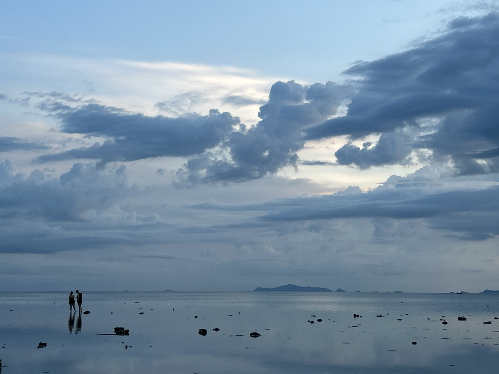The image depicts a tranquil seascape at dusk or dawn on Koh Phangan, Thailand. Two silhouetted figures, possibly a couple, stand in shallow water near the shore, their reflections shimmering on the calm, mirror-like surface. Scattered dark objects, likely rocks or debris, dot the water. In the distance, a chain of islands is visible against the horizon, silhouetted against a dramatic sky filled with a mix of soft, light clouds and darker, more voluminous formations. The overall mood is one of peace and serenity, with the soft, diffused light suggesting a time of day when the sun is low. The sky's color palette ranges from a pale blue at the top to softer, muted tones as it approaches the horizon.