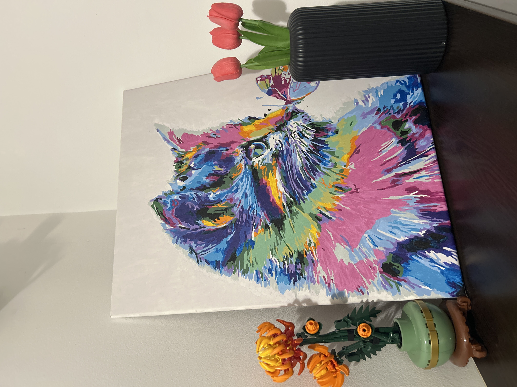 The scene depicts a wall with a painting leaning against it, flanked by floral decorations. The painting is an abstract representation of an animal, possibly a dog or cat, rendered in a variety of colors including pink, blue, purple, yellow, and green. A butterfly is painted on the animal's head. Above the painting is a black rectangular object and three red tulips. Below the painting is a Lego flower. The wall is painted white. The surface underneath the right corner of the painting is dark brown. The location is Unknown, Unknown.