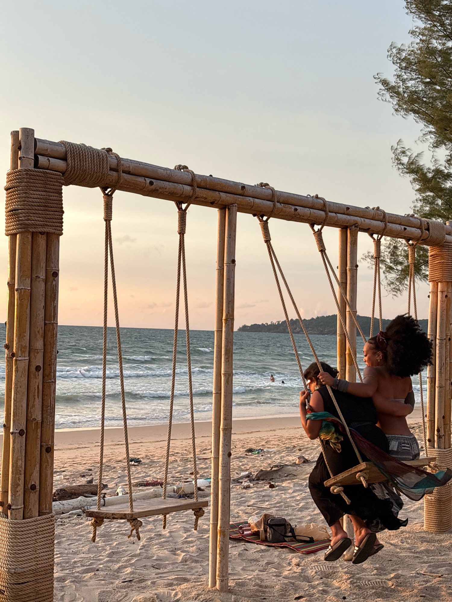 In Choeng Thale, Thailand, two individuals are captured on a beach at dusk, enjoying a bamboo swing set. The scene depicts a serene coastal environment with gentle waves rolling onto the sandy shore, and a hint of the sun setting on the horizon, casting a warm glow. The swing itself is a rustic construction of bamboo poles and ropes, supporting a wooden seat. One person, on the right, has dark, voluminous curly hair and is facing away from the camera, embracing the other individual. The second person, on the left, is positioned slightly behind and to the side, facing towards the ocean. They are both dressed in casual beachwear. The ground beneath the swing is covered with sand, with a small mat and a bag placed nearby. The background reveals the vast ocean stretching to a distant coastline dotted with hills. The overall atmosphere is one of relaxed enjoyment and connection amidst a beautiful natural setting.
