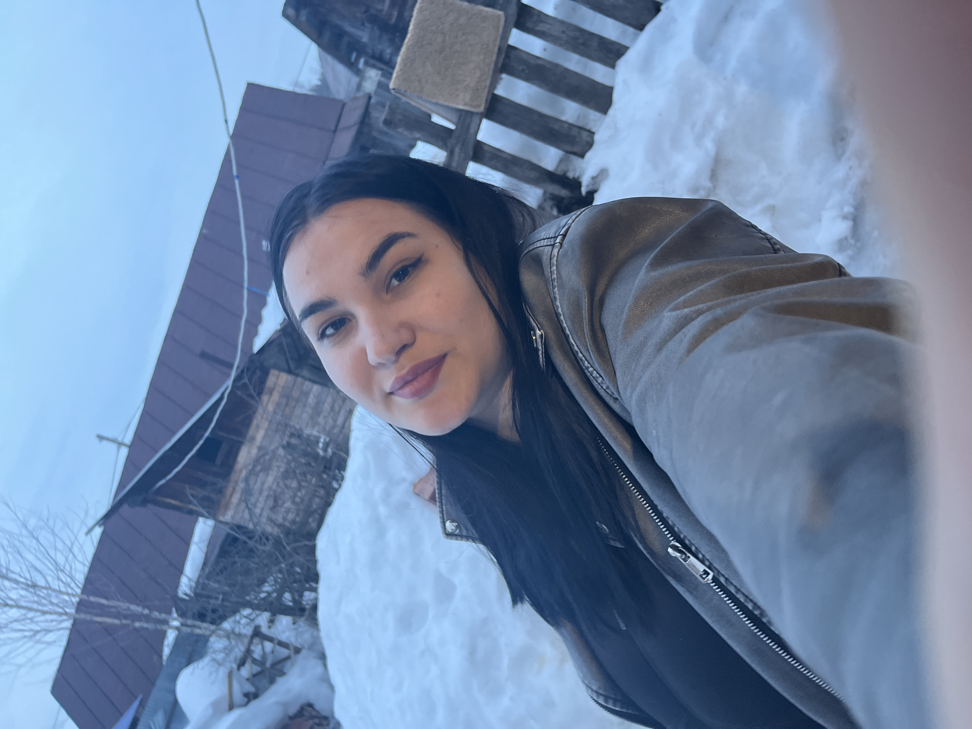 A female individual is depicted in a close-up selfie, captured outdoors in the city of Chaikovsky, Russia. She is positioned in the foreground, angled slightly to the left, looking directly at the camera with a subtle, closed-mouth smile. She has dark hair, dark eyebrows, and is wearing a greyish-brown or olive-toned jacket with a visible front zipper and collar studs.
The background shows a landscape extensively covered in snow, indicating a winter or early spring setting. To the left and center-left, a wooden building with a dark brown corrugated metal roof is visible, partially obscured by snowdrifts and the branches of a bare deciduous tree. To the right of the subject, a rustic wooden plank fence stands, with a light-colored rectangular object, resembling a folded textile or mat, draped over its top rail. Further in the distance on the right, additional structures with dark roofs and snow-covered ground are discernible. The sky above is overcast and light blue-grey.