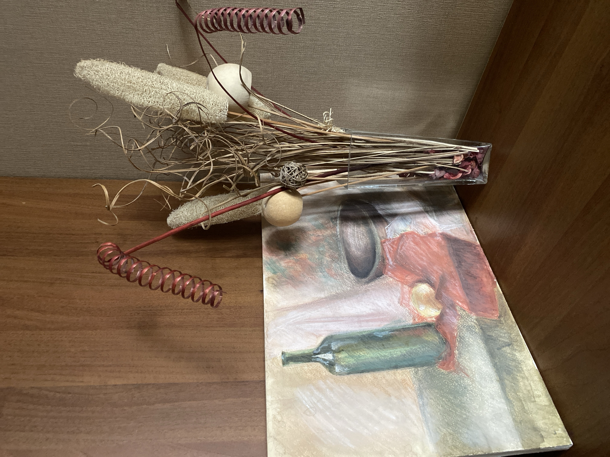 The image presents a still life arrangement on a wooden surface, possibly a table or shelf. The backdrop consists of a plain wall with two different shades of neutral color. The composition features a vase filled with dried floral elements, including thin sticks, curled tendrils, and seed pods. The vase is a clear, rectangular glass shape containing what appear to be dried petals. In the foreground, partially overlapping the still life, is a painting depicting a bottle and other objects. The arrangement is likely indoors, and while there is nothing in the image to confirm it is located in Izhevsk, Russia, the arrangement itself doesn’t provide any evidence against the possible location.