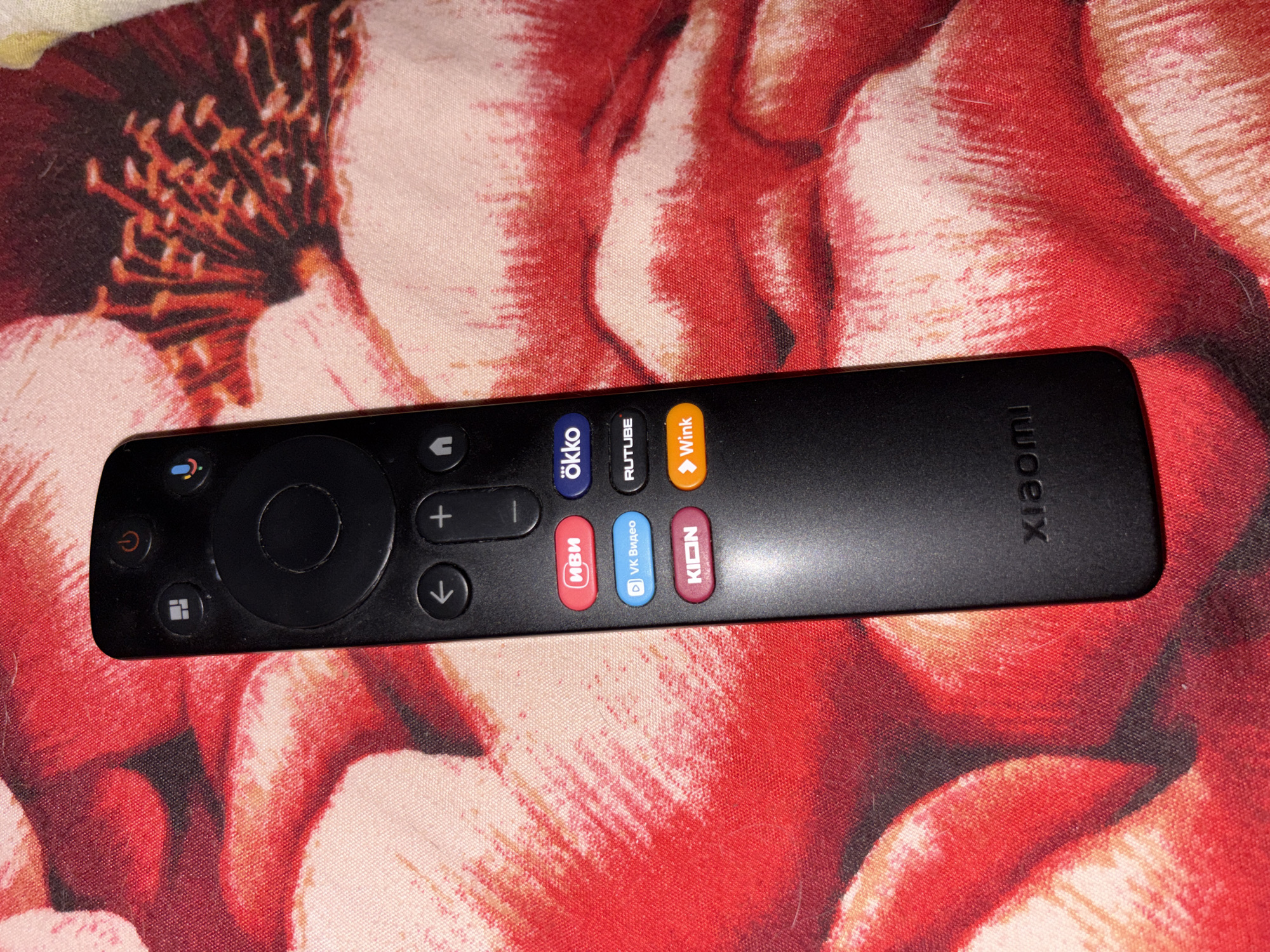 A black Xiaomi remote control is placed on a red and pink floral patterned fabric. The remote has several buttons, including a power button, a microphone button, navigation buttons, and shortcut buttons for applications like IVI, OKKO, VK Video, Rutube, KION, and Wink. The fabric appears to be a tablecloth or blanket. The image does not depict any specific location within the city of Засечное, Russia.