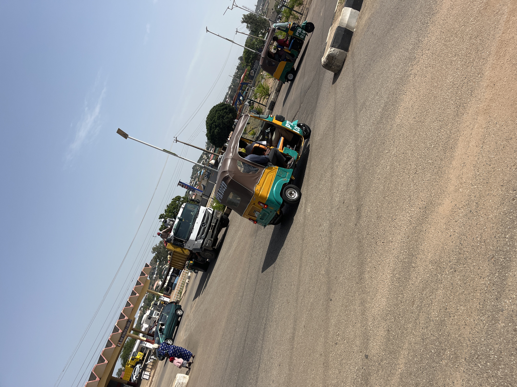 The image shows a street scene in Bukuru, Nigeria. Two auto-rickshaws, also known as Keke Napeps, are traveling down a paved road. The auto-rickshaws are yellow and green and appear to be carrying passengers. One auto-rickshaw has "PTA 059" visible on its side. A white truck with a yellow cargo bed is also present, with people in the back. A green car is parked on the side of the road. Further down the road, a pedestrian overpass with signage is visible, and beyond that, buildings and trees suggest a populated area. The sky is clear and blue, indicating it is daytime, likely late morning or early afternoon given the bright sunlight casting shadows. The overall impression is of a busy street with daily transportation activities.