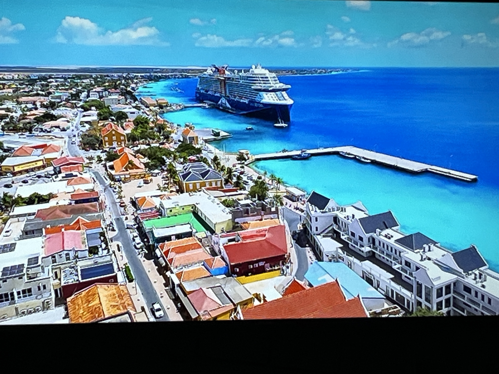 The image depicts an aerial view of a coastal city with brightly colored buildings and a large cruise ship docked in the harbor. The city features a grid-like street layout with numerous houses and commercial buildings, many with orange, red, and green rooftops. Palm trees are visible, suggesting a tropical or subtropical climate. The ocean is a vibrant turquoise color, and a long pier extends into the water. A large cruise ship, predominantly blue and white, is moored next to the pier. The sky is clear with a few scattered white clouds. The scene is tranquil and picturesque, portraying a lively tourist destination. The provided image and context do not suggest any connection to the city of Kharkiv, Ukraine, as it is geographically impossible for it to have a coastal setting and cruise ships.