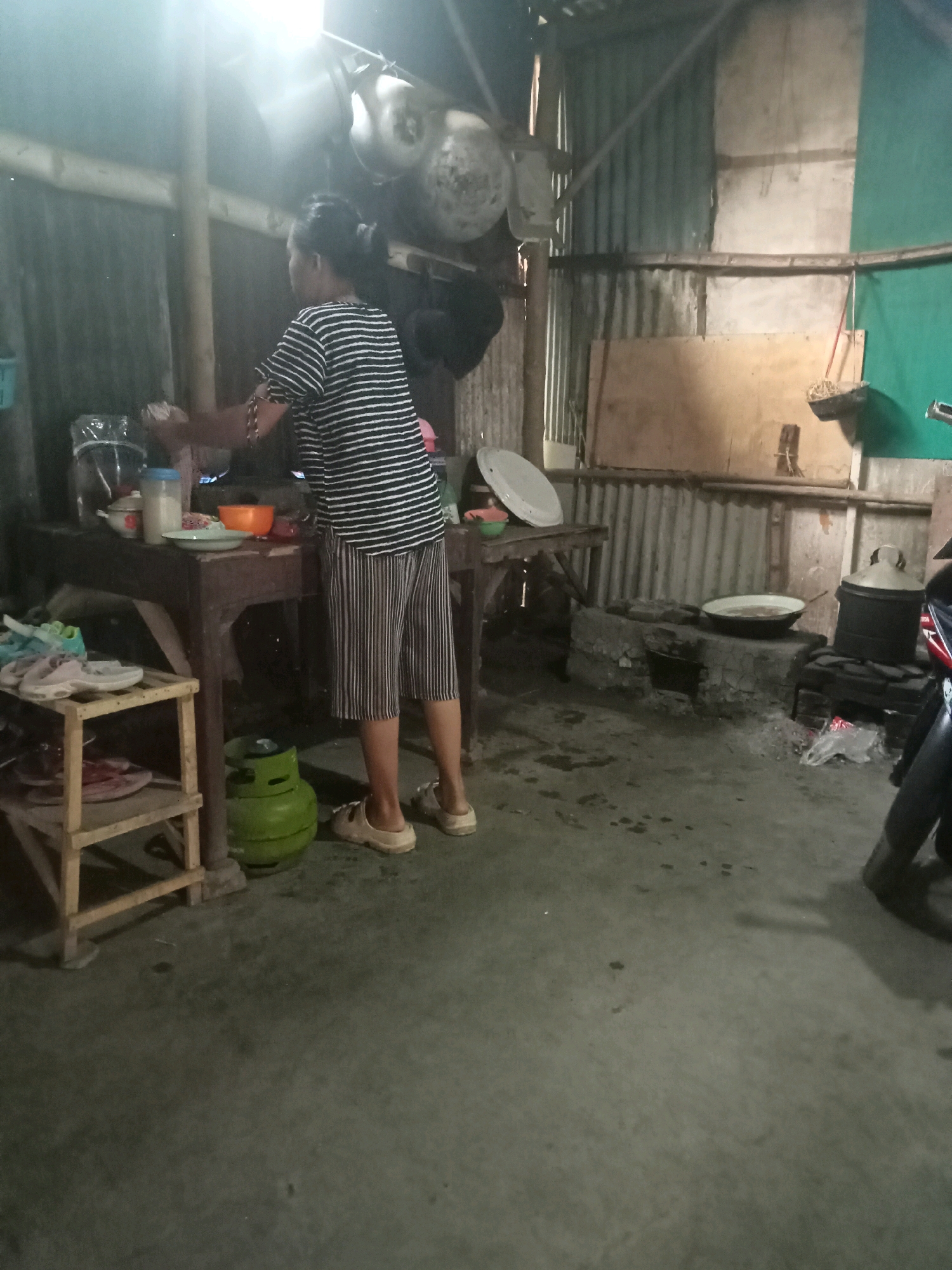 A woman stands in a dimly lit, rustic kitchen, preparing food at a wooden table. She wears a striped t-shirt and striped pants, with her hair pulled back. Various kitchen items, including bowls, a gas cylinder, and shoes on a wooden shelf, are visible. The kitchen features corrugated metal walls, a traditional stove, and a motorcycle parked in the background. The scene appears to be set in a modest home in Brumbungan Kidul, Indonesia.