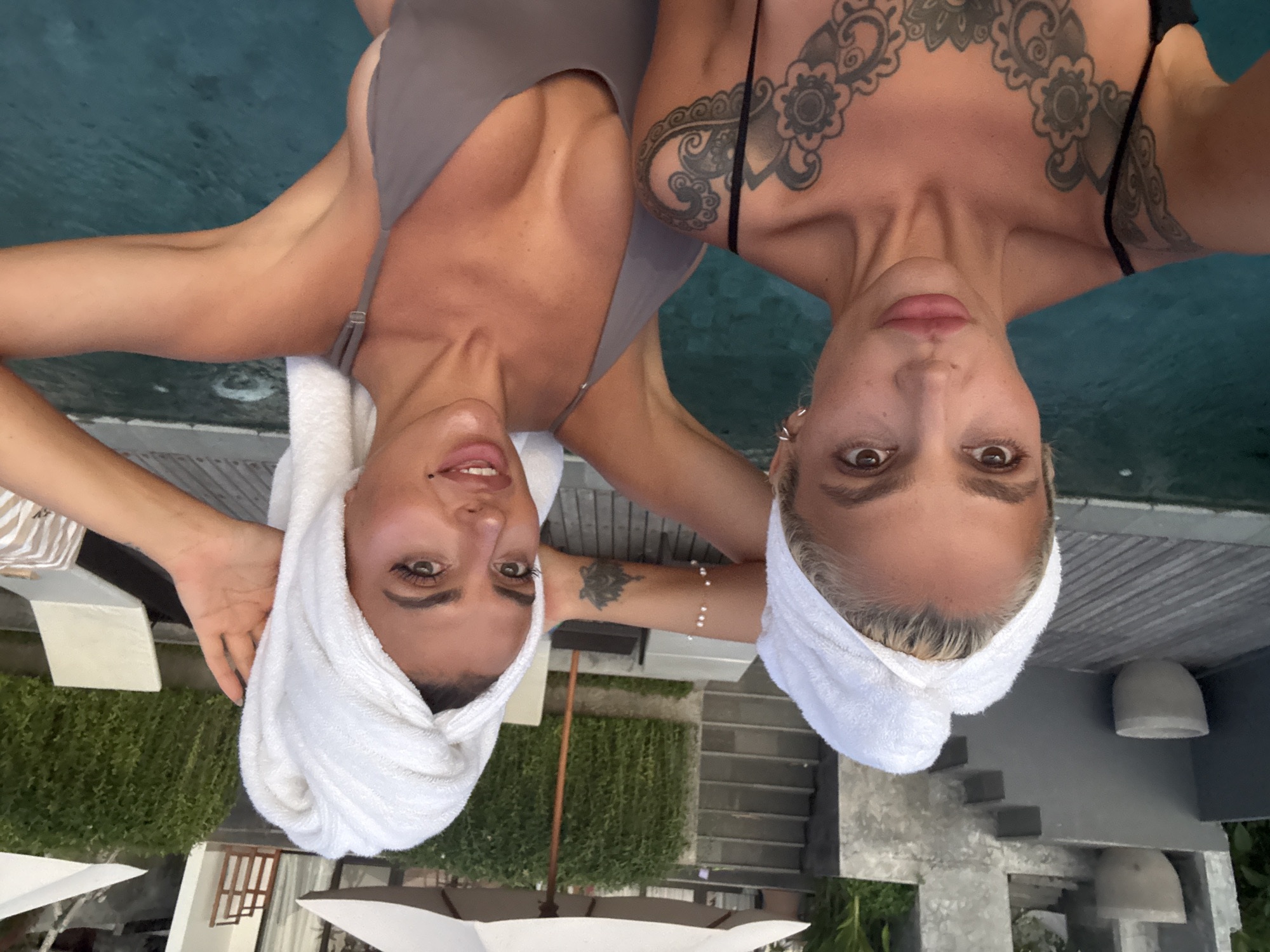 Two women, wearing towels on their heads and swimwear, pose for a selfie by a swimming pool. The woman on the left has blonde hair and extensive tattoos on her chest and shoulder. The woman on the right has dark hair and is wearing a grey swimsuit. The background shows lush greenery, lounge chairs, umbrellas, and a stone staircase, consistent with a resort in Kuta Selatan, Indonesia. The lighting suggests it is late afternoon or early evening.