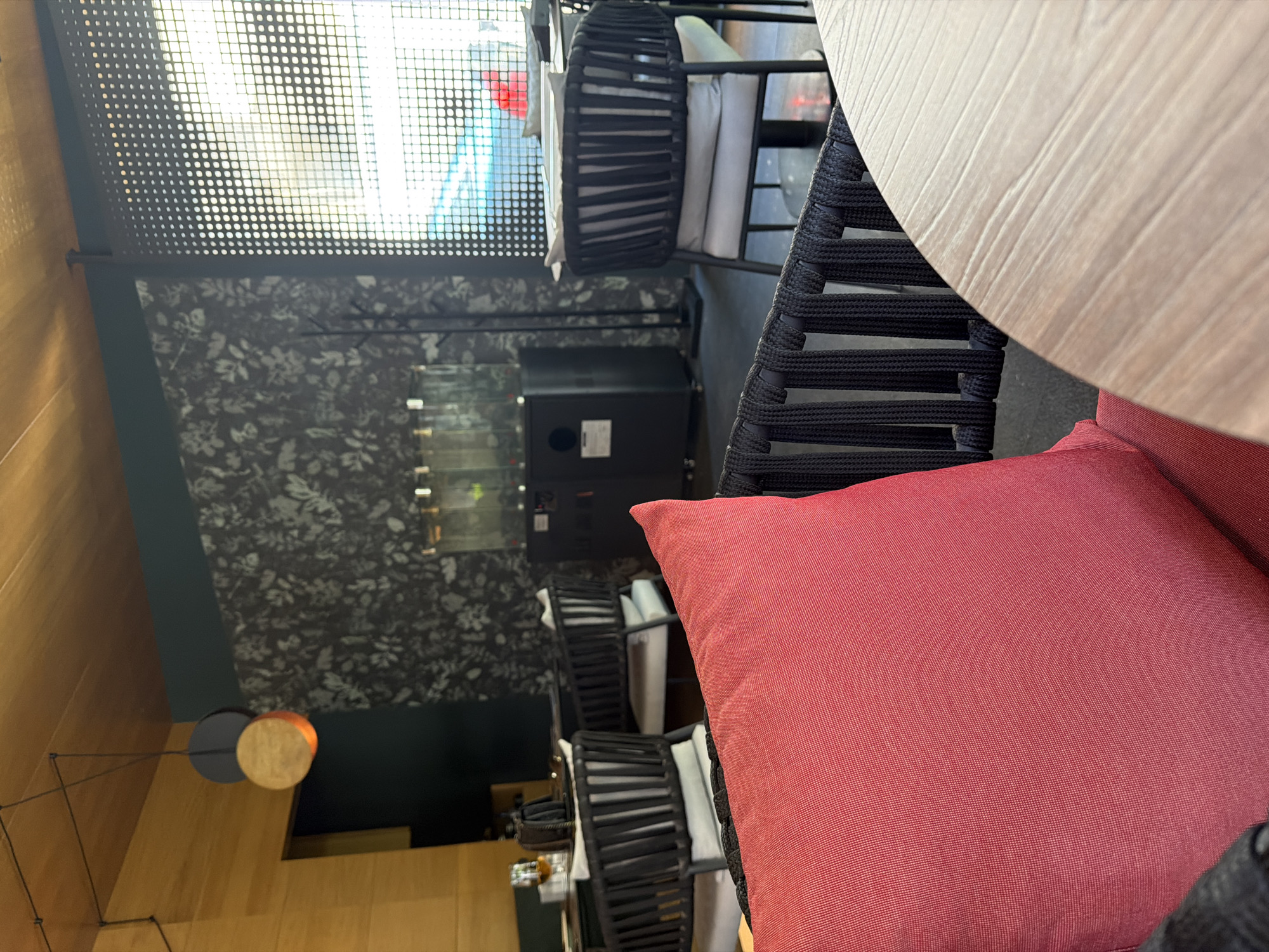 The image shows a restaurant interior in Kazanlak, Bulgaria. A table with a light-colored, wooden-style top is partially visible in the upper right. A black woven chair with a red cushion is in the foreground. The chair faces an open space featuring a black metal screen in front of a window. Below the screen, a wall with a gray floral design is visible, along with a black cabinet or display case. A modern, suspended lighting fixture with a circular lampshade is on the left wall. There are additional black woven chairs with white cushions farther back in the space. The interior has a warm and modern aesthetic.