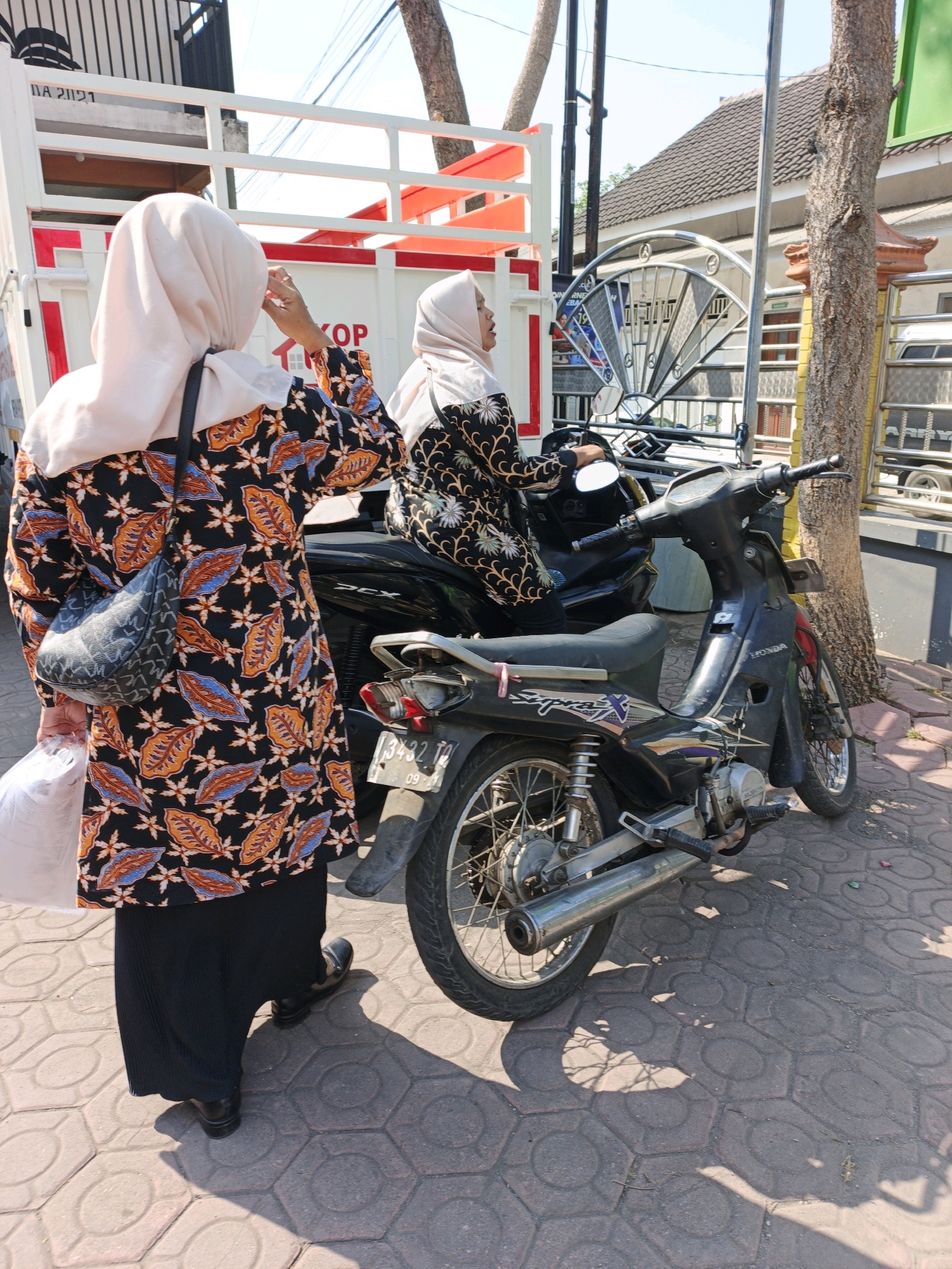 In the city of Ketegan, Indonesia, a photograph captures two women standing near a motorcycle on a sun-drenched sidewalk. The woman on the left, facing away from the camera, wears a light pink hijab and a black batik-patterned top with orange and blue leaf motifs. She also wears a black pleated skirt and black shoes. A dark patterned purse hangs over her left shoulder. She is holding a white plastic bag in her left hand and appears to be adjusting her hijab with her right hand. Next to her, slightly in front, a second woman, also wearing a light pink hijab, is seated on a black motorcycle. She is looking to her right, towards the left of the frame, and is wearing a black top with a floral pattern. The motorcycle is a black Honda Supra X with a license plate that reads "3432 TD 09-24". The sidewalk is paved with hexagonal concrete blocks, and shadows from the surroundings are cast across it. Behind the women, a white truck with a red-and-white striped railing is visible, and further in the background, there are buildings with tiled roofs, a tree with rough bark, and a decorative metal gate. The scene suggests a moment captured in a street or market area.