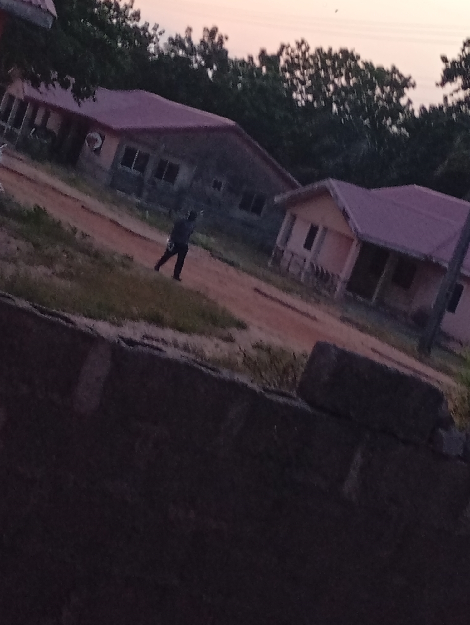 The image shows a person walking on a dirt road between two buildings in Unknown, Unknown. The buildings have red roofs and light-colored walls. The sky is dusky. A low brick wall is in the foreground.