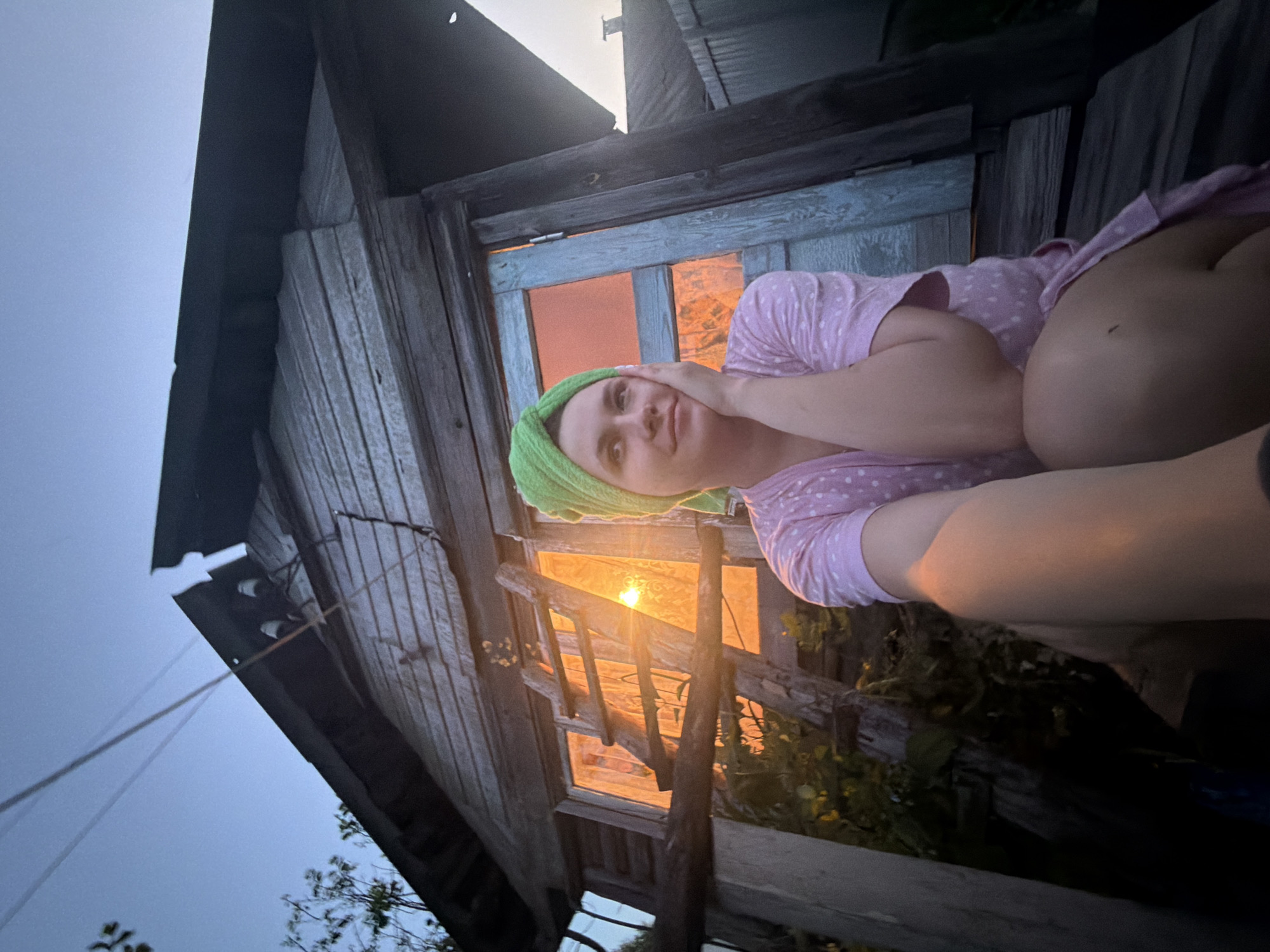 In the village of Krasny Mayak, Russia, a young woman is photographed outdoors in front of a dilapidated wooden building. She is wearing a pink polka-dot robe and a green towel wrapped around her head. Her left hand is resting on her cheek, and she is looking to her left. The setting appears to be dusk, with the sky a pale blue and the sun's rays casting an orange glow through a window in the building. A wooden ladder leans against the exterior of the structure. The overall scene suggests a rustic, possibly rural, environment.