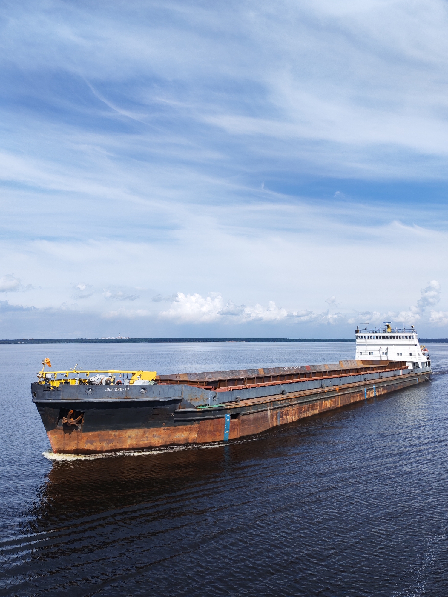 A heavy barge, labeled "ПЕВСКИИ-13", travels on a wide, dark body of water under a bright, cloudy sky. The barge is long and rectangular, with a rusty brown cargo area and a white superstructure at the stern. The bow of the barge is dark gray with an anchor visible. The water is calm with gentle ripples. In the distance, a dark green tree line separates the water from a pale sky. The image appears to have been taken from a higher vantage point, possibly another vessel or a bridge. The exact location in Kostyaevka, Russia cannot be determined from the visual information provided.