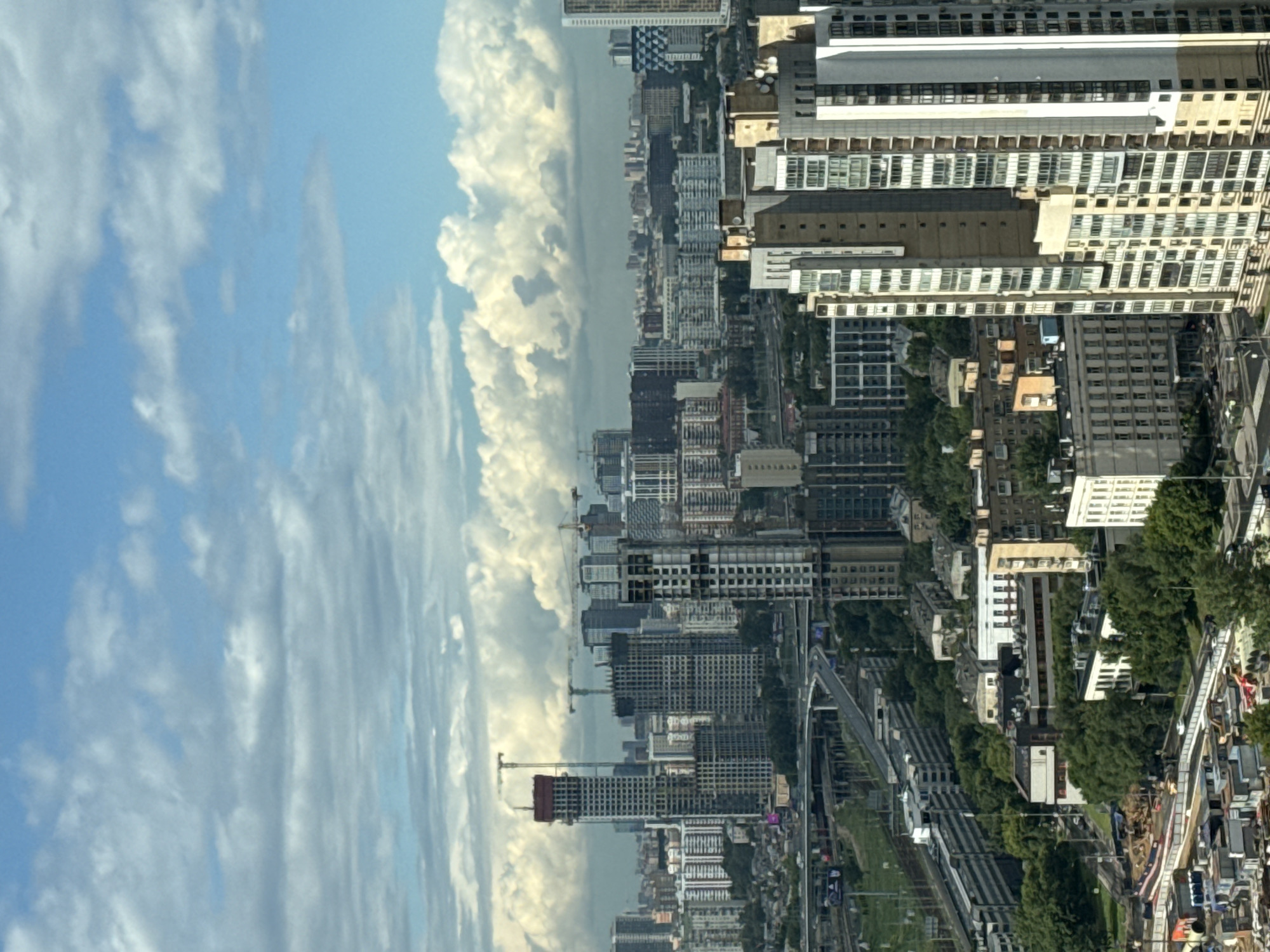 The media file is an aerial, sideways view of a city skyline in Khimki, Russia, with a large, cloudy sky on the left. The city features a mix of completed and under-construction buildings, including tall residential towers and industrial structures. Cranes are visible amidst the construction, indicating ongoing development.
The scene is characterized by a bright, daytime setting with a sky filled with fluffy, white cumulus clouds against a pale blue background. The city itself is densely packed with buildings of varying architectural styles and heights, many with numerous windows. Some buildings appear to be residential complexes with a uniform design, while others are more varied. Green trees are interspersed throughout the urban landscape, particularly in the lower right portion of the image.
Below the skyline, there are visible transportation networks, including what appear to be railway tracks or elevated roadways. A road with some traffic is also discernible in the lower right corner, with a few people and vehicles. The overall impression is one of a developing urban area with a busy atmosphere under a pleasant sky. No visible text is present in the image.