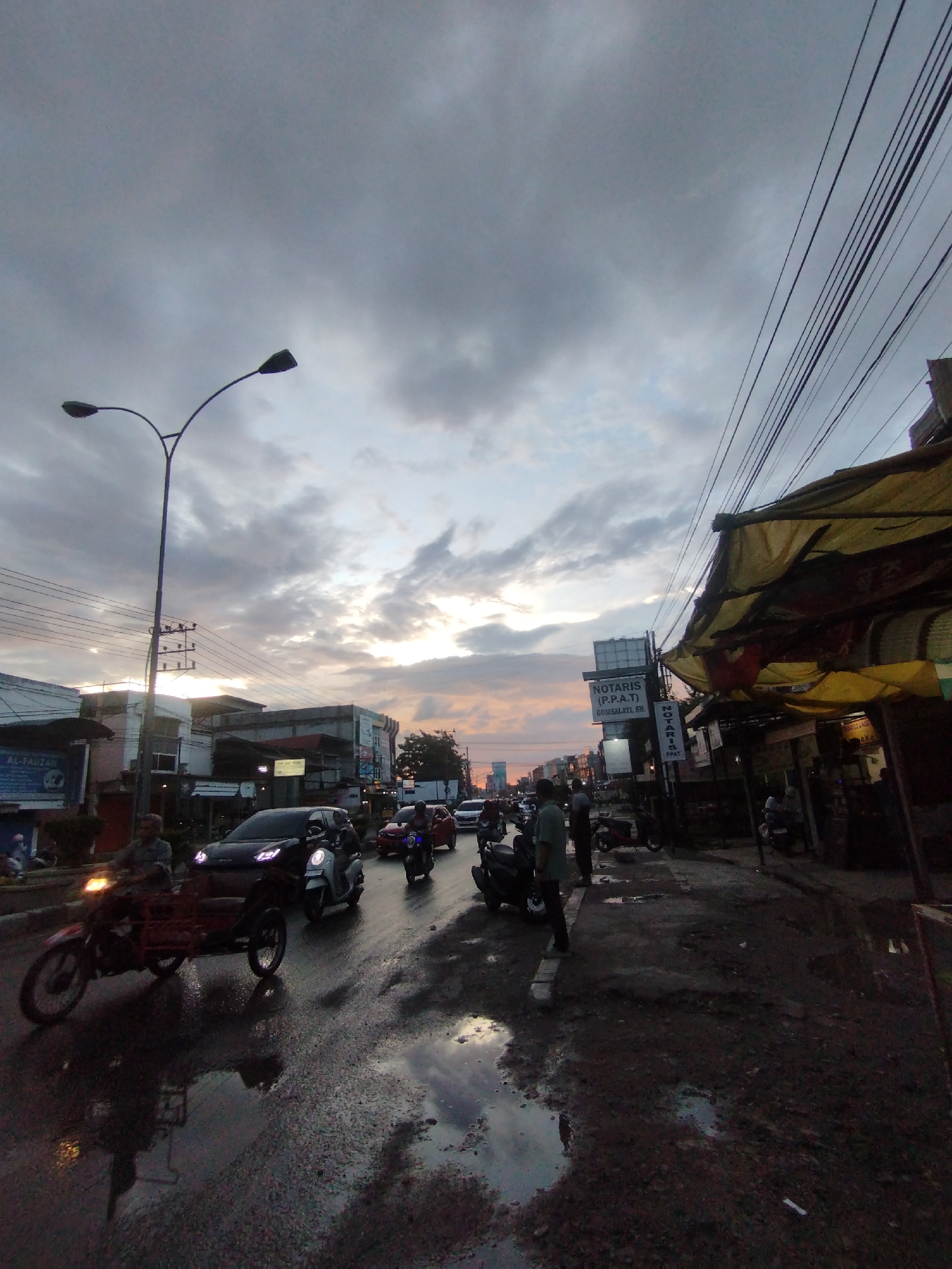 The image depicts a street in Lhokseumawe, Indonesia, under an overcast sky at dusk. The street is wet, reflecting light. Several vehicles are on the road, including a motorcycle with a sidecar, a scooter, a car, and other motorcycles. People are standing on the sidewalk near shops with awnings. A street lamp is visible. Power lines crisscross above. Signage in Indonesian is present, including "NOTARIS (P.P.A.T)". The street surface is uneven and shows signs of disrepair.