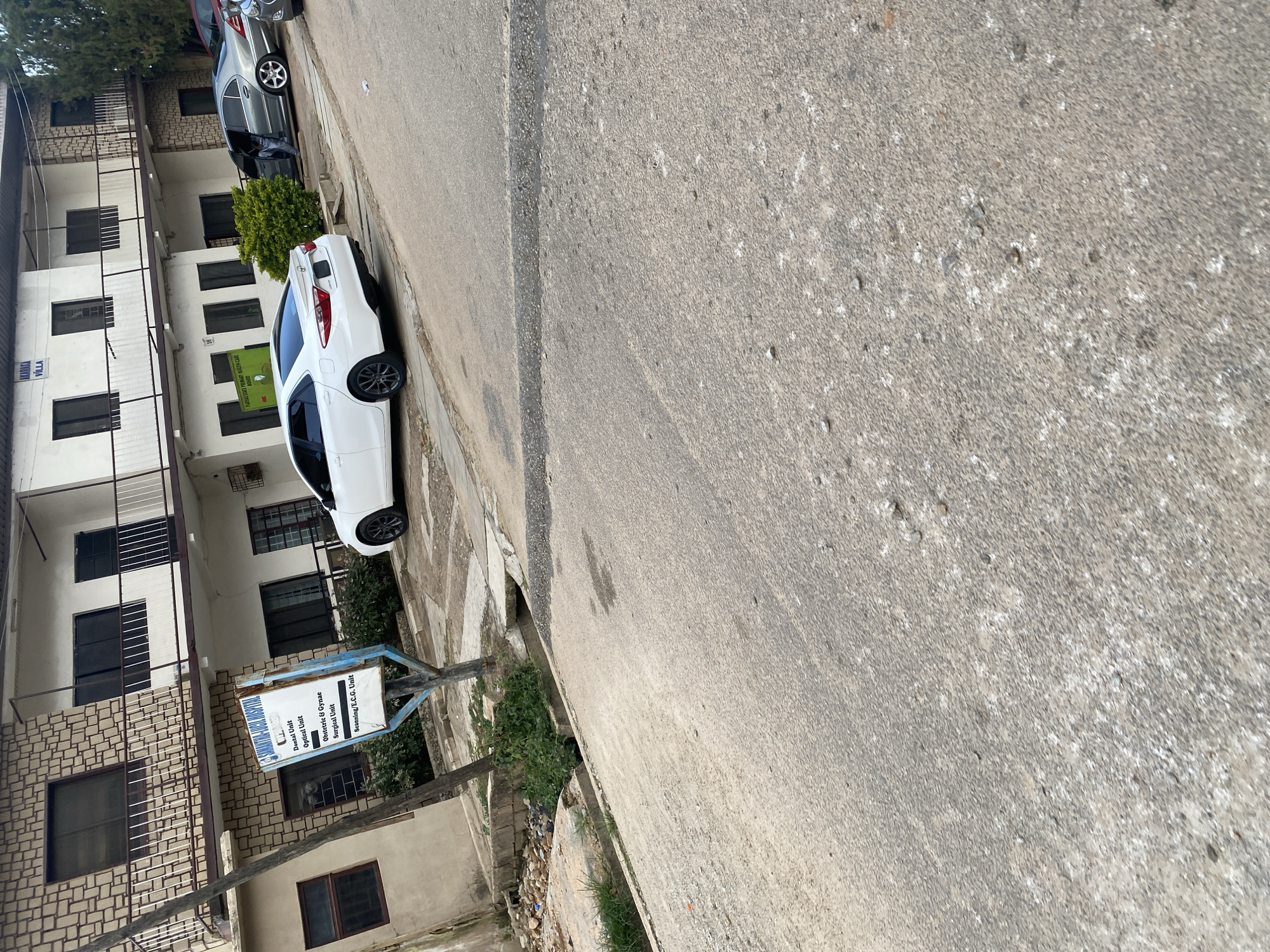 A full-shot, eye-level outdoor image captures a street scene in Jos, Nigeria, on a slightly overcast day. The primary focus is a white sedan parked on the right side of the frame. A grey sedan is parked behind it, with its driver's side door open and a person partially visible inside. In the background, a multi-story building with a rough, textured facade and white painted sections is visible. A signpost in front of the building displays hospital services. The foreground and midground are dominated by a paved road and sidewalk, with scattered debris and vegetation along the edges. The scene appears to be a commercial or residential area.