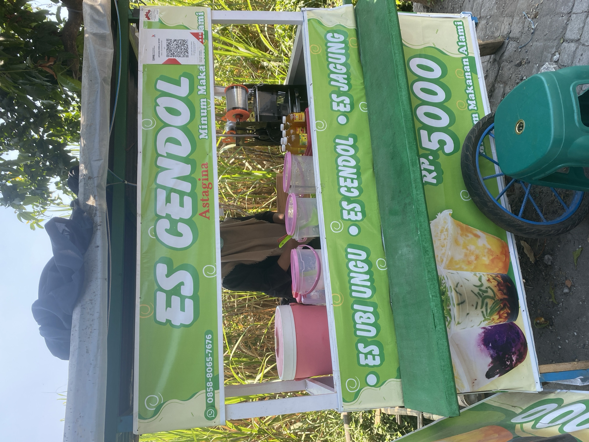The image shows a street food cart selling "Es Cendol" and other drinks in the city of Kediri, Indonesia. The cart is brightly decorated with green and white signage. Prominently displayed on the cart are the words "ES CENDOL," "ES UBI UNGU," and "ES JAGUNG." A price of "RP. 5000" is also visible. Behind the counter, a person wearing a hijab is seen preparing drinks. Various containers and bottles are organized on the cart. The cart is positioned outdoors, with tall green plants in the background, suggesting a rural or semi-rural setting within Kediri City. A green plastic stool and a spare tire are present near the cart. The overall scene depicts a typical roadside food vendor operation in Indonesia.