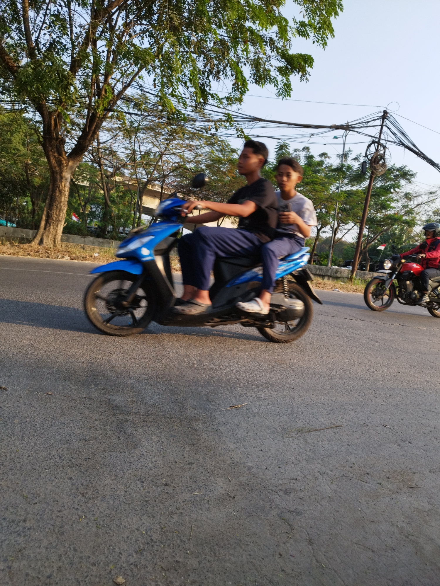 Two young males are riding a blue scooter on a paved road. The driver, in a black t-shirt, is in the front, and a passenger, wearing a light-colored t-shirt, is behind him. They are both wearing casual clothing. The scooter has a visible front fender, wheel, and part of the engine. The road surface appears to be asphalt. In the background, there are trees with green leaves and some electrical wires suspended overhead. To the right, another motorcycle is visible with a rider wearing a red jacket and a helmet. The lighting suggests it is daytime, possibly late afternoon, with shadows cast on the road. The environment appears to be an urban or suburban setting with vegetation and infrastructure.