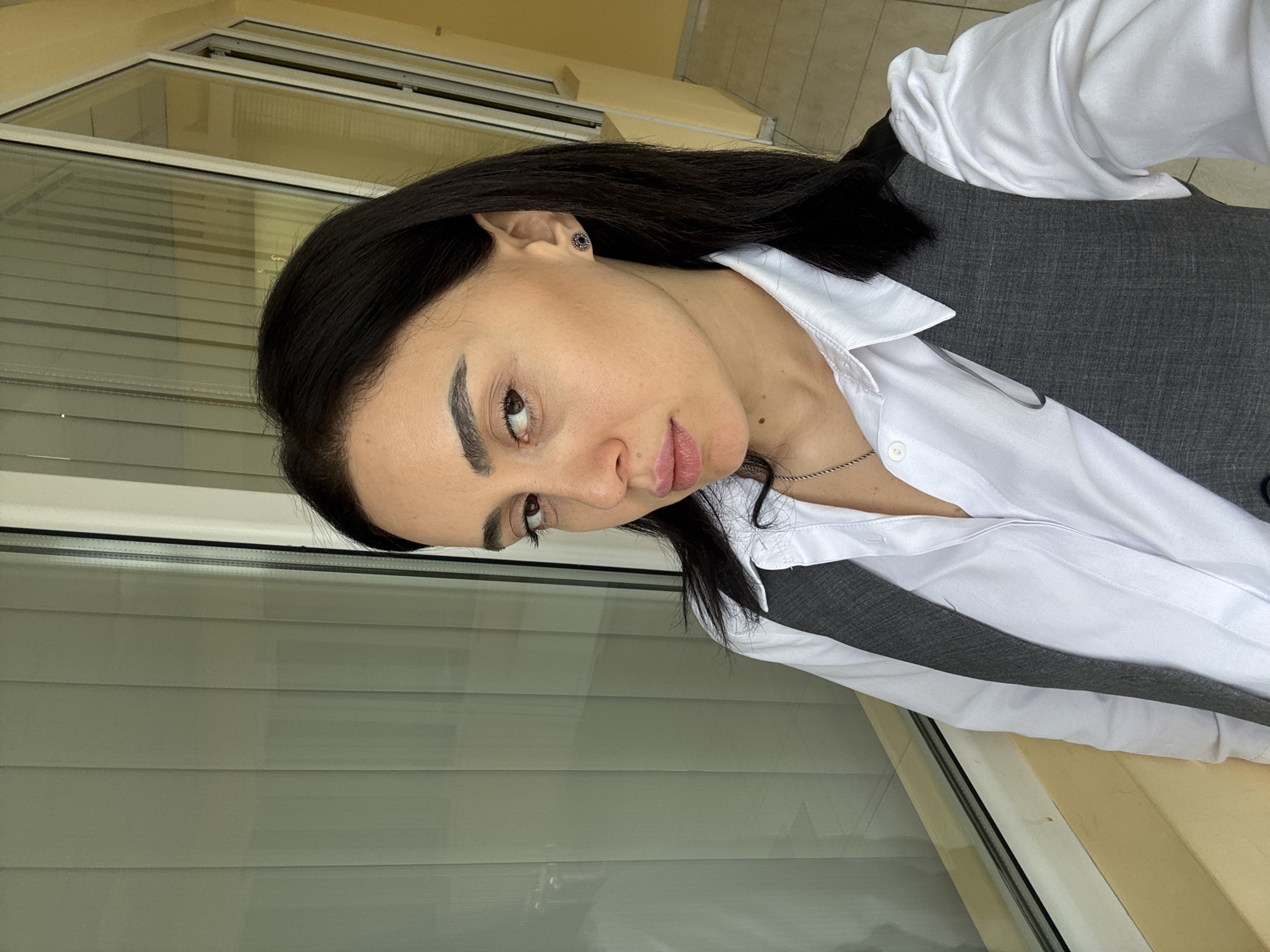 A woman with dark hair is photographed in a rotated close-up shot. She is wearing a white button-down shirt and a gray vest. The background appears to be a window with blinds. The location seems to be a balcony or terrace setting with a view of a building, likely in the city of Tbilisi, Georgia, based on the architectural style potentially visible through the window.