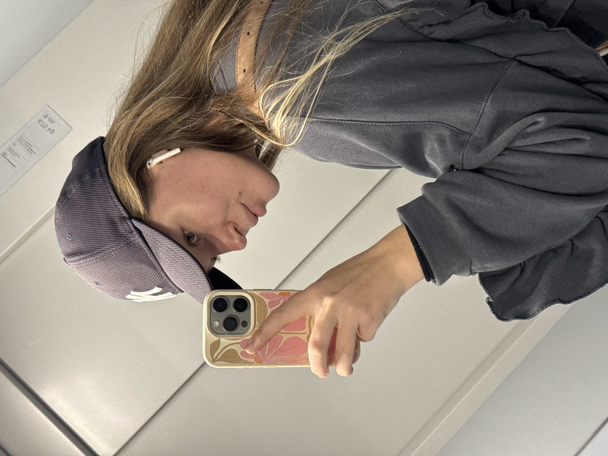A woman with blonde hair and a grey baseball cap is taking a selfie in a mirror. She is wearing a grey sweatshirt and has an earbud in her left ear. She is holding a phone with a floral case. The background is a plain white wall. The location cannot be determined as Vozdovac Urban Municipality, Serbia based on the image alone.