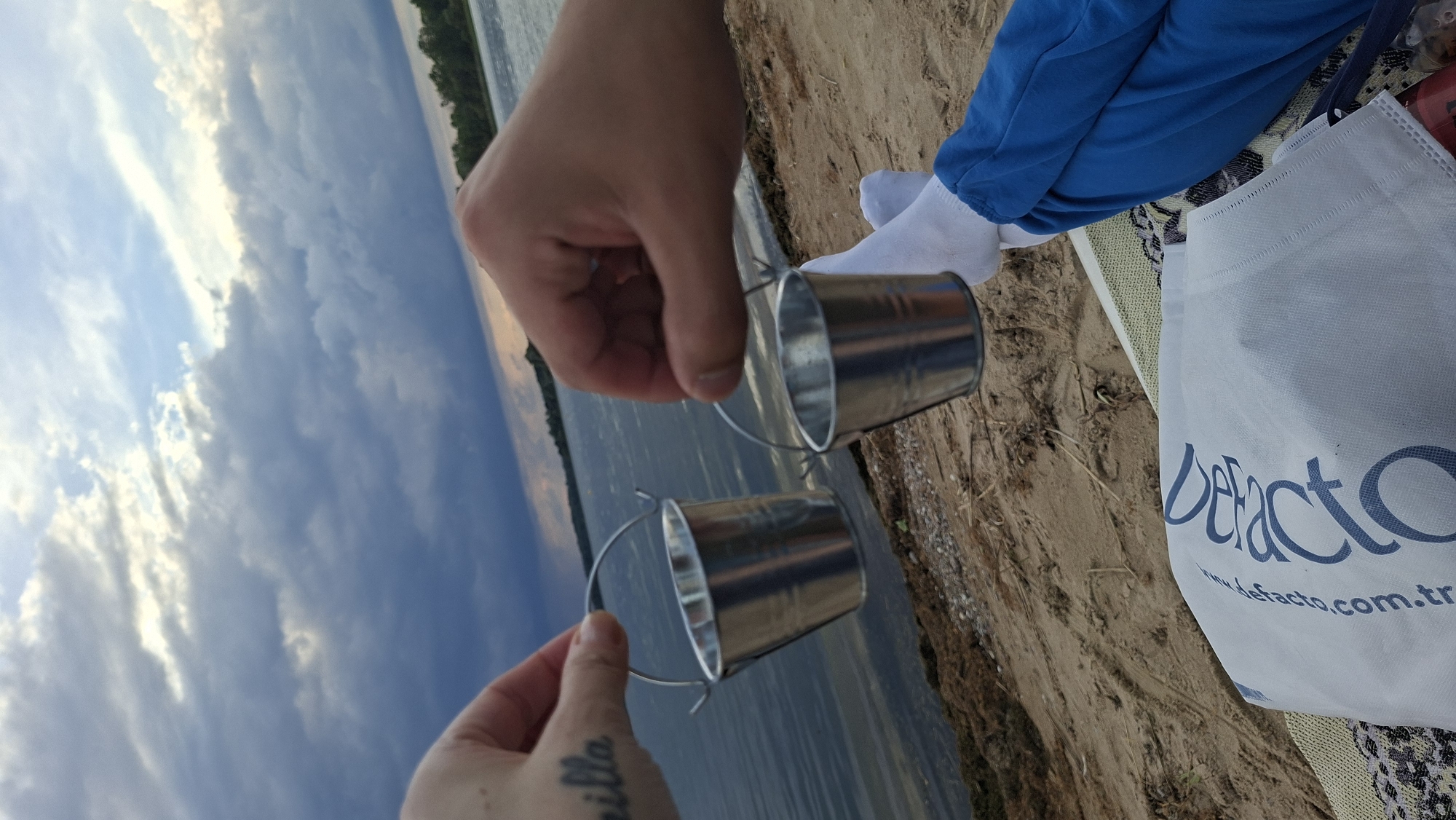 The image depicts a close-up of two hands holding small, silver buckets over a body of water. The person on the left has a tattoo on their arm, partially visible. The background features a cloudy sky and a body of water with a tree line in the distance. In the foreground, there is a sandy beach and a white tote bag with blue lettering. The scene appears to be set at a lake or river. The location is identified as the city of Myadzyel, Belarus.
