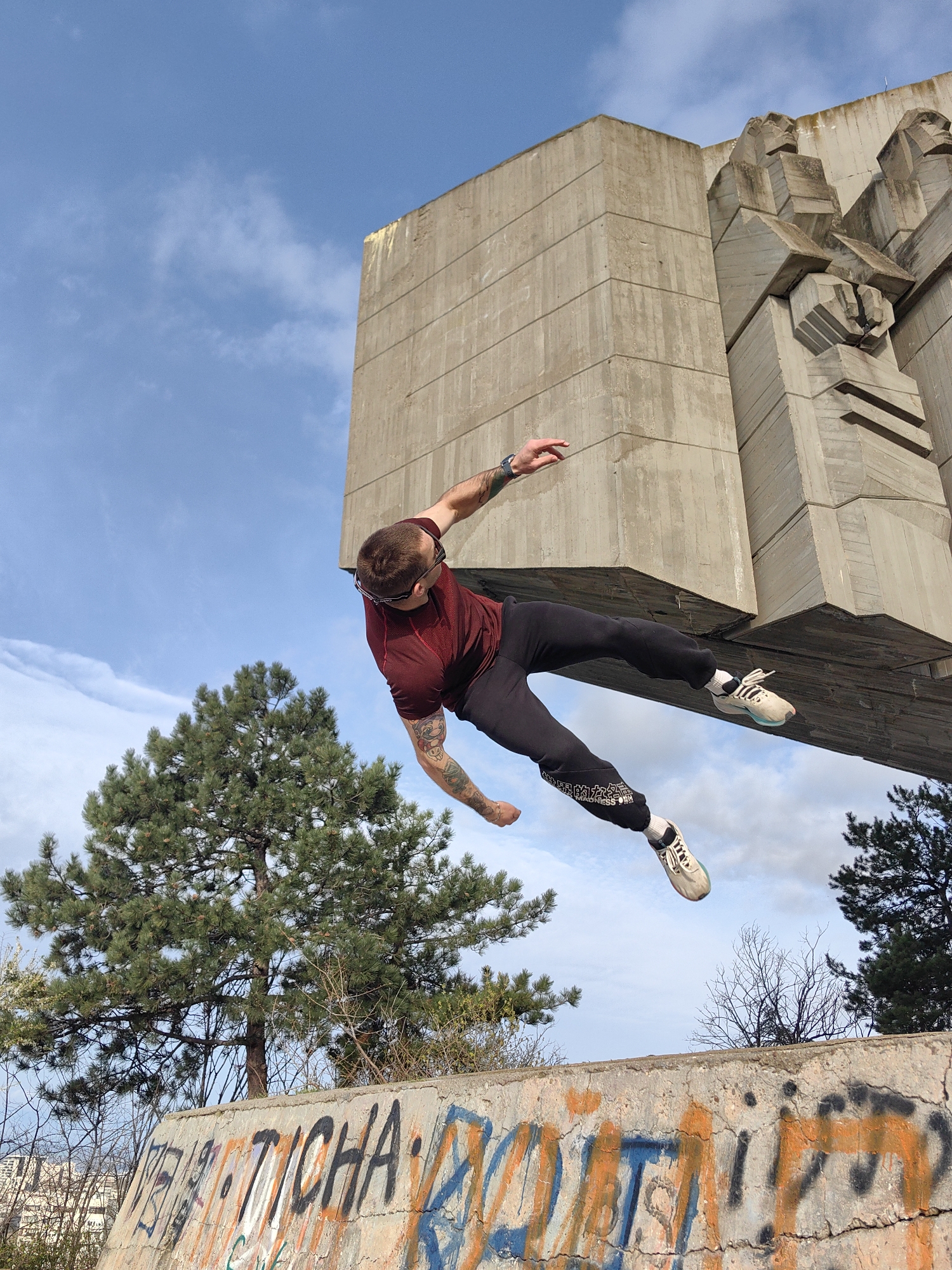 A man in mid-air is performing a parkour maneuver next to the Monument of the Bulgarian-Soviet Friendship in Varna, Bulgaria. He is wearing a maroon shirt, black pants with white lettering, and white sneakers with turquoise soles. His body is angled horizontally. He has a tattoo on his left arm and a watch on his left wrist. The monument is made of large concrete blocks, and a tree is visible to the left. The man is jumping over a concrete wall covered in graffiti. The sky is blue with scattered clouds.