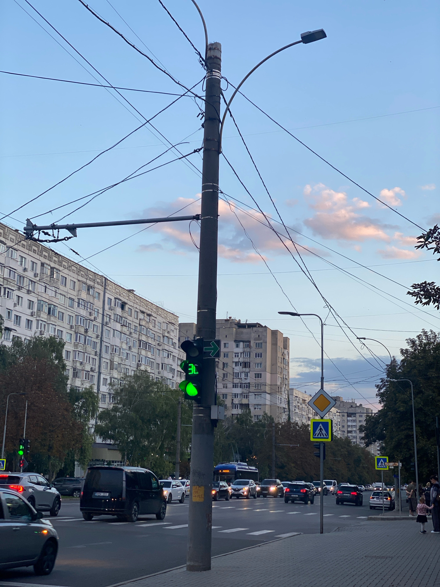 The image captures a street scene in Chișinău, Moldova, likely during the late afternoon or early evening, given the soft light and pastel-colored clouds. A prominent traffic light, showing a green arrow and a green circle, is visible on the right side of the frame, next to a utility pole adorned with numerous overhead wires. In the background, a large, multi-story apartment building with many windows stretches across the left side of the image. Other buildings, including a taller, lighter-colored structure, are visible further in the distance. A busy street with multiple lanes of traffic flows through the scene, with cars, vans, and a bus visible. Pedestrians are present on the sidewalk to the right, with a couple of people standing near a crosswalk. Traffic signs, including a yellow diamond-shaped sign indicating a main road and a blue square sign with a white pedestrian symbol, are positioned at the intersection. Trees and foliage line the sides of the street, and overhead streetlights are also visible. The sky is a clear blue with scattered pink and white clouds.