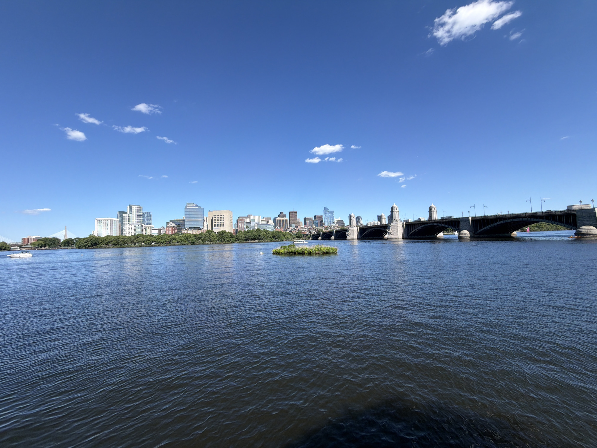 This is a wide, horizontal, eye-level shot of a city skyline across a body of water. The sky is a clear, bright blue with a few wispy white clouds. In the foreground, dark blue water fills the bottom half of the frame, with small ripples on its surface. A small, circular patch of green vegetation, appearing to be an island, floats in the middle of the water.
Beyond the water, a city skyline stretches across the horizon. The buildings vary in height and architectural style, with a mix of modern skyscrapers and older structures. A large, arched bridge with stone towers spans the water to the right of the frame. Several boats are visible on the water, including a white motorboat on the left and a pink object on the water near the bridge. The location is Cambridge, United States of America.