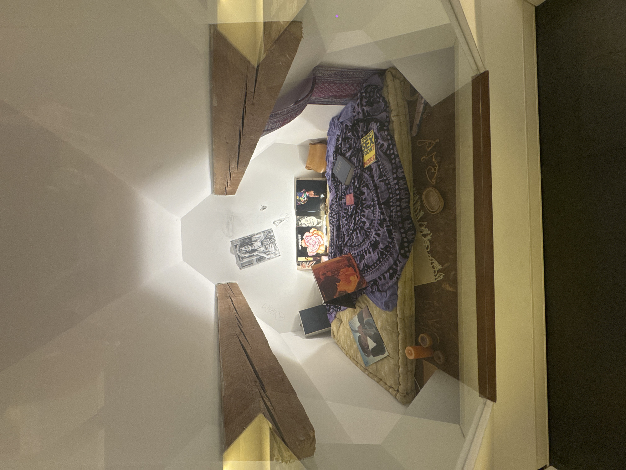 The image displays a staged representation of a bohemian-style room, possibly a recreated historical exhibit. The room features a mattress on the floor with a purple patterned blanket draped over it. Various objects are scattered on and around the mattress, including records, books, a purse, and candles. The space is framed by slanted wooden beams and white walls, suggesting an attic or loft environment. Pictures, including one resembling Janis Joplin, are affixed to the walls. The scene appears to be enclosed in a glass display, indicating it is a preserved or recreated setting. The context of this image has no connection to the city of Bern, Switzerland.