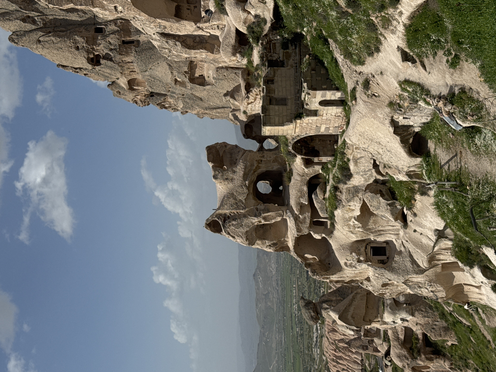 The image depicts a landscape in Uçhisar Beldesi, Turkey, characterized by eroded rock formations and cave dwellings. The sky is partly cloudy, with blue patches visible. In the distance, a valley with agricultural fields is visible. The rock formations feature numerous holes and openings, suggesting they are either natural caves or man-made dwellings carved into the soft rock. Some structures appear to be more substantial, possibly representing older buildings or settlements. Patches of grass and greenery are visible along the top of the formations.