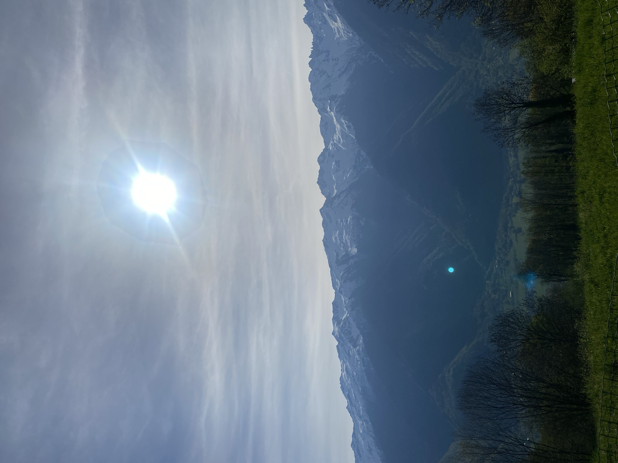 The image presents a vibrant, sunlit mountain landscape, captured in Saint-Georges-d'Hurtières, France. The sky, dominating the left side of the frame, is bright blue with delicate, wispy white clouds stretching across it. A very intense, overexposed sun, surrounded by prominent lens flares and a sunburst effect, is positioned in the upper left quadrant, casting a strong light across the scene.
A majestic range of snow-capped mountains runs horizontally across the middle of the image. The sun illuminates their jagged peaks, highlighting the white snow, while their lower slopes and the deep valleys below them appear in stark, dark blue shadows.
In the foreground, towards the right, a darker, forested valley is visible. Bare trees, typical of early spring or late autumn, are silhouetted against the shadowed slopes, though some patches of green foliage can be discerned. On the far right, a strip of bright green grass, possibly a field or pasture, is bordered by a wire fence. Several distinct blue circular lens flares are visible in the dark valley area, reflections of the bright sun.
The scene depicts a clear, sunny daytime, likely morning or late afternoon, given the strong sun and deep shadows. There are no people or specific activities depicted. No visible text is present in the image.